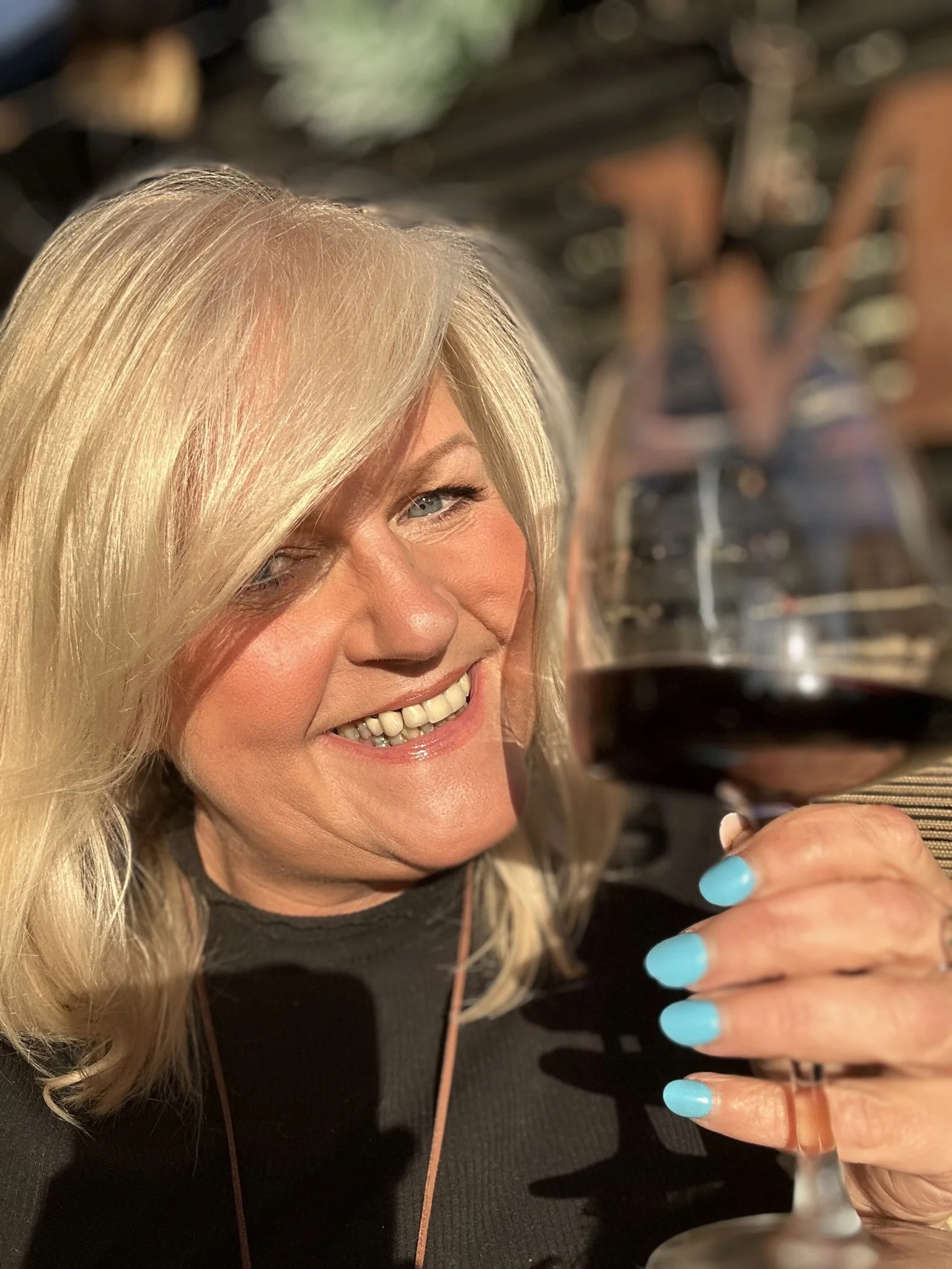 Person with blonde hair holding a glass of red wine, smiling, with bright blue nail polish, in a sunny outdoor setting.