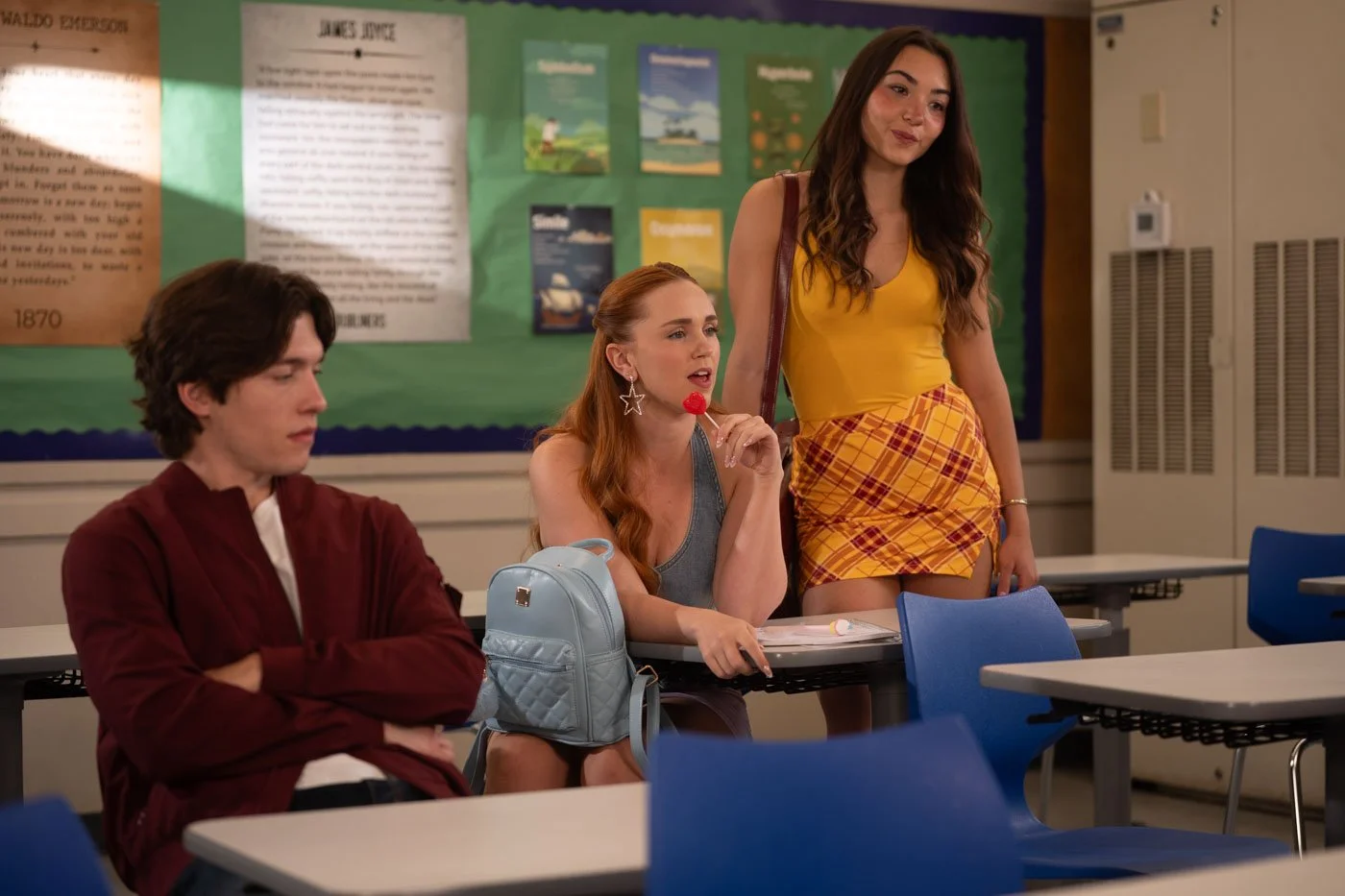 A classroom scene with three young women; one sitting at a desk with a blue backpack, one standing, and another sitting with a lollipop, all engaged in conversation. Green bulletin board in background.
