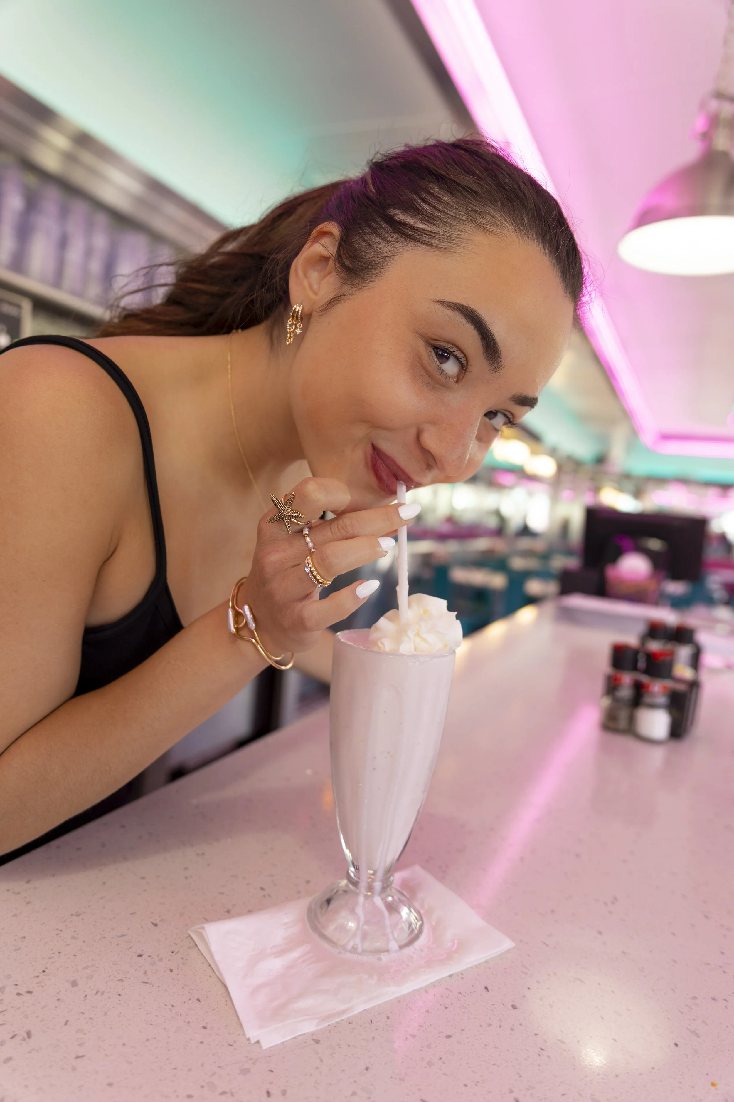 A young woman with dark hair in a ponytail, wearing a black sleeveless top and gold jewelry, is sitting at a pink countertop in a diner. She is sipping a milkshake from a tall glass with a straw, whipped cream topping, wearing star-shaped earrings an
