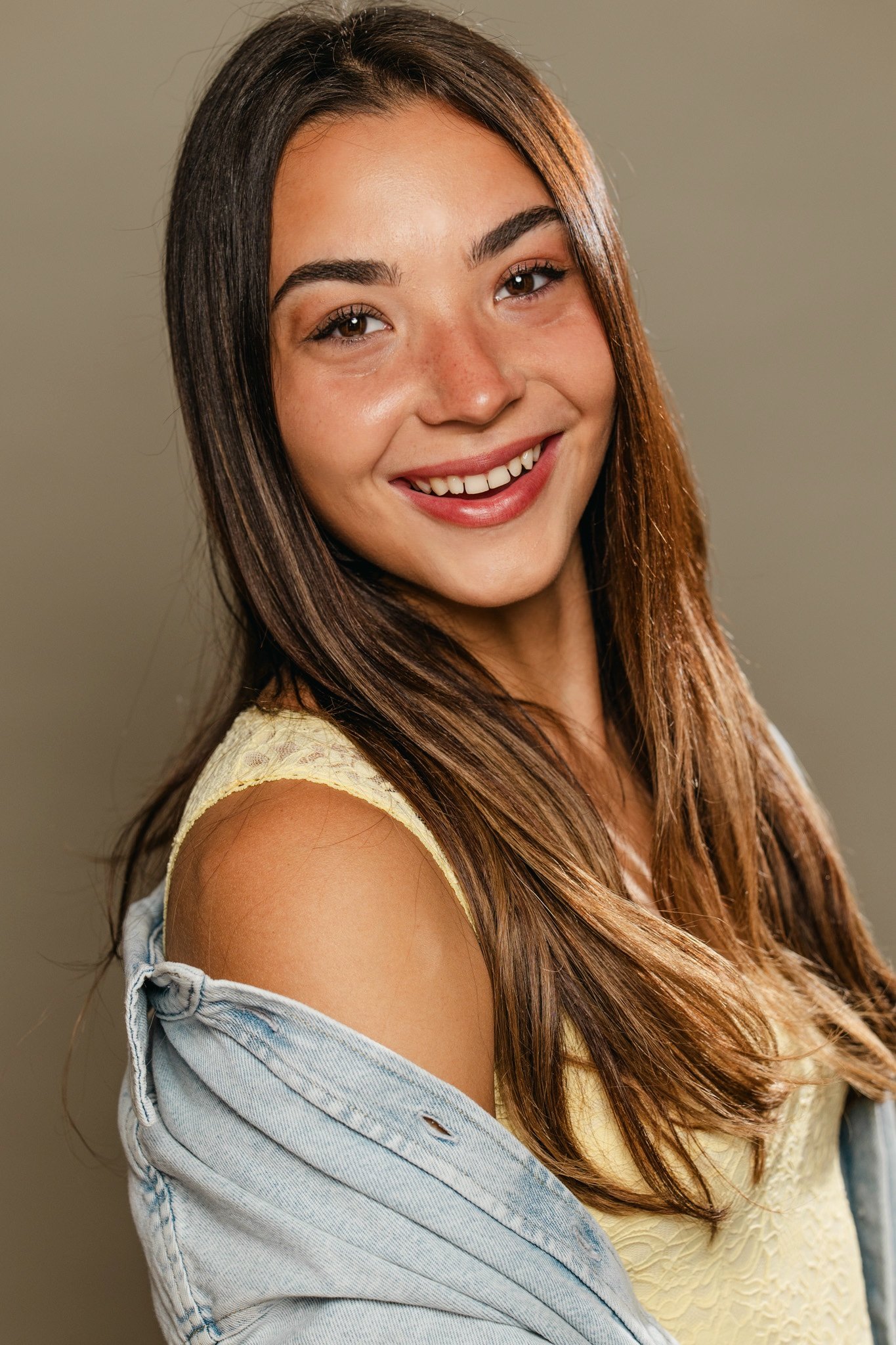 A young woman with long brown hair smiling, wearing a yellow tank top with lace, and a light blue denim shirt off her shoulder.