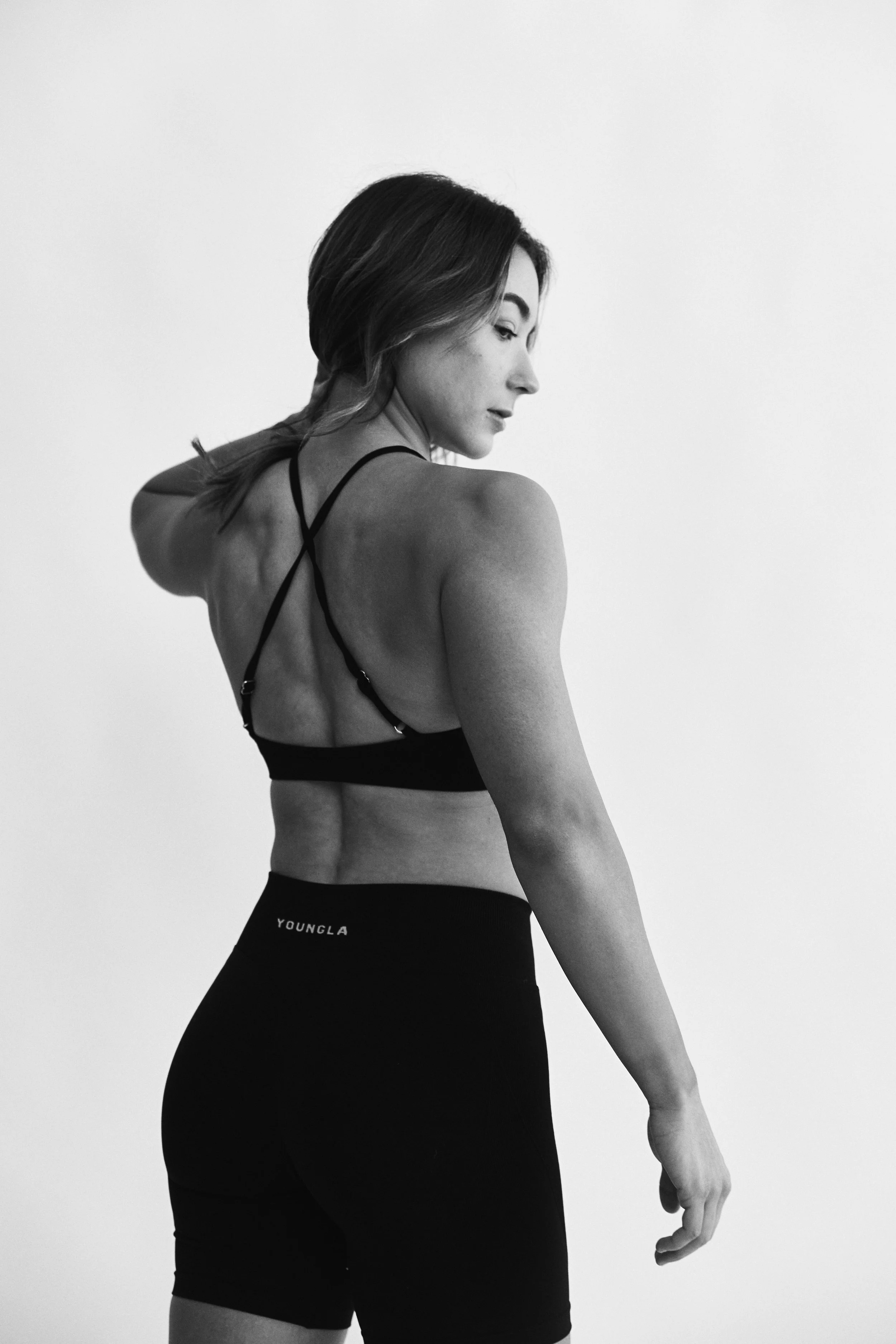 Black and white photo of a woman with short hair, wearing a sports bra and high-waisted shorts, looking down and to the side in a minimalistic setting.