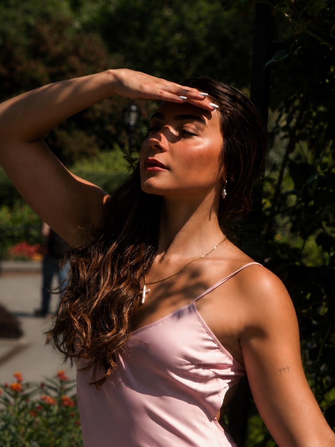 A woman with long, wavy brown hair wearing a light pink satin spaghetti strap top, jewelry including a necklace with a cross pendant and earrings, standing outdoors in sunlight with her hand on her forehead, against a background of green trees and flowers.