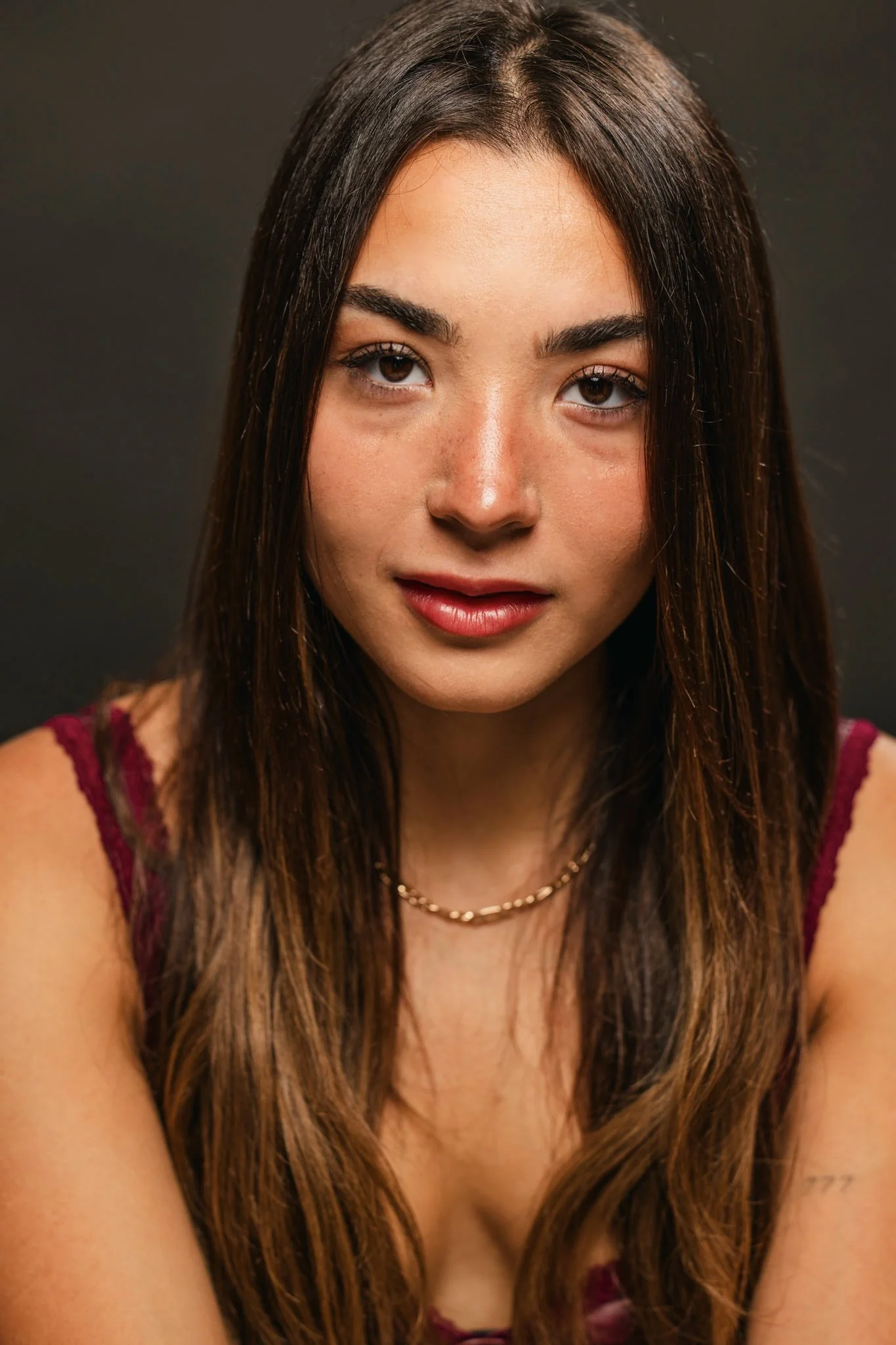 Close-up portrait of a young woman with long brown hair, wearing a gold chain necklace and a red top, against a dark background.