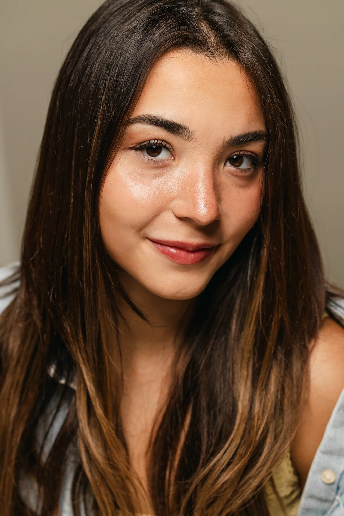 Close-up of a woman with long, brown hair and brown eyes, smiling softly, wearing a light-colored top and a denim jacket.