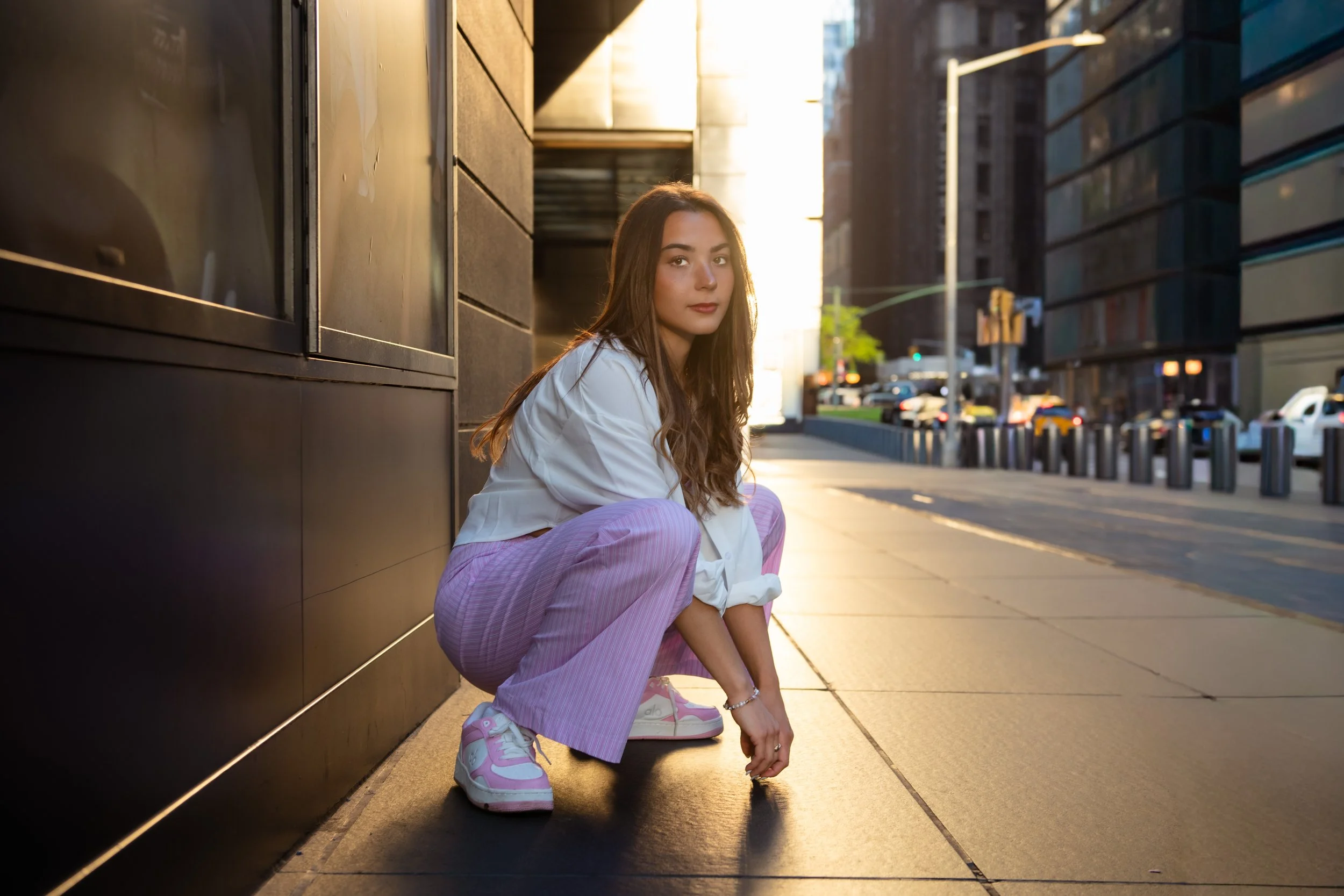 Young woman crouching on the sidewalk in an urban setting during sunset, wearing a white jacket, pink striped pants, and pink sneakers, with buildings and cars in the background.