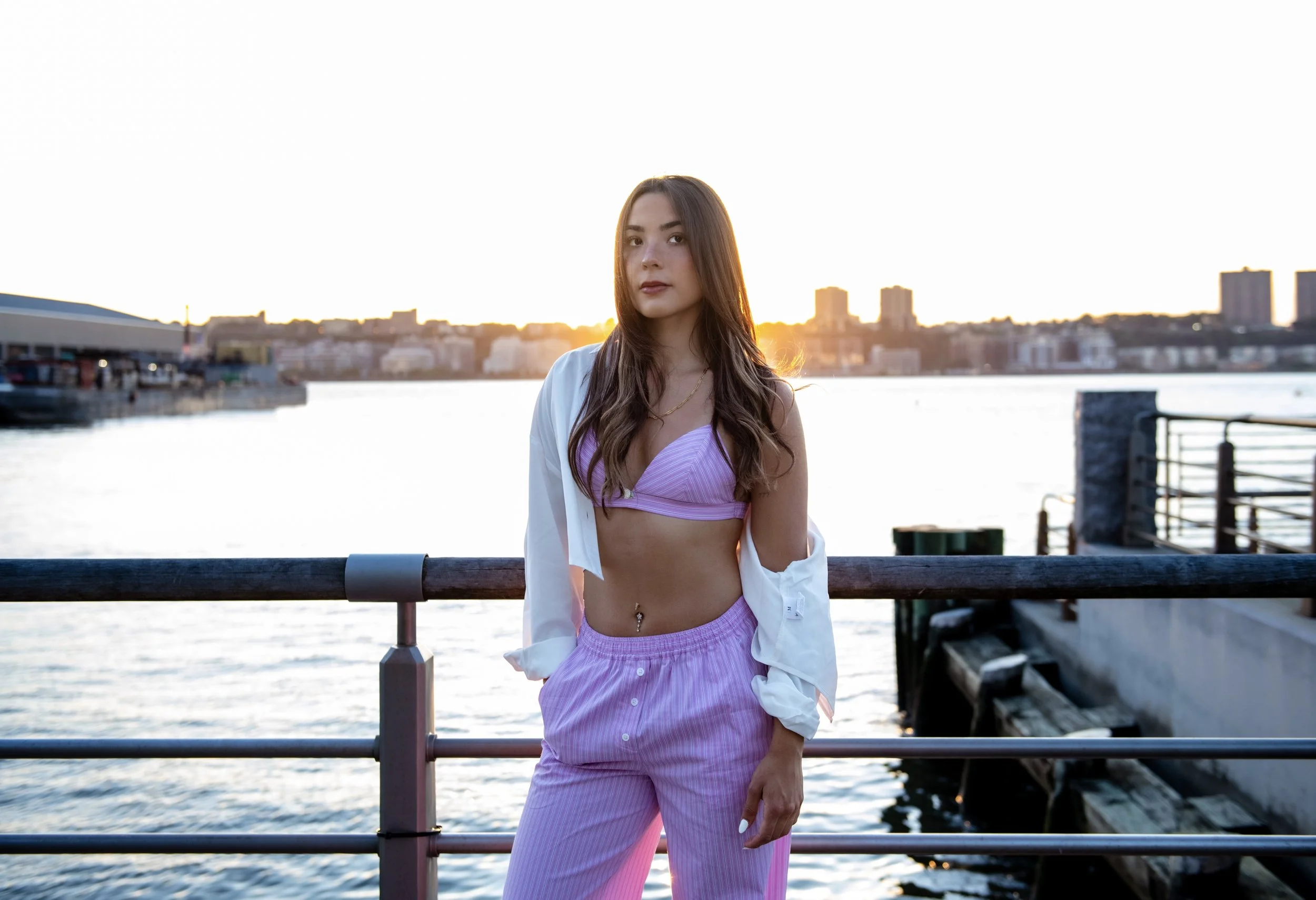 A young woman in a pink outfit standing by a waterfront during sunset with a city skyline in the background.