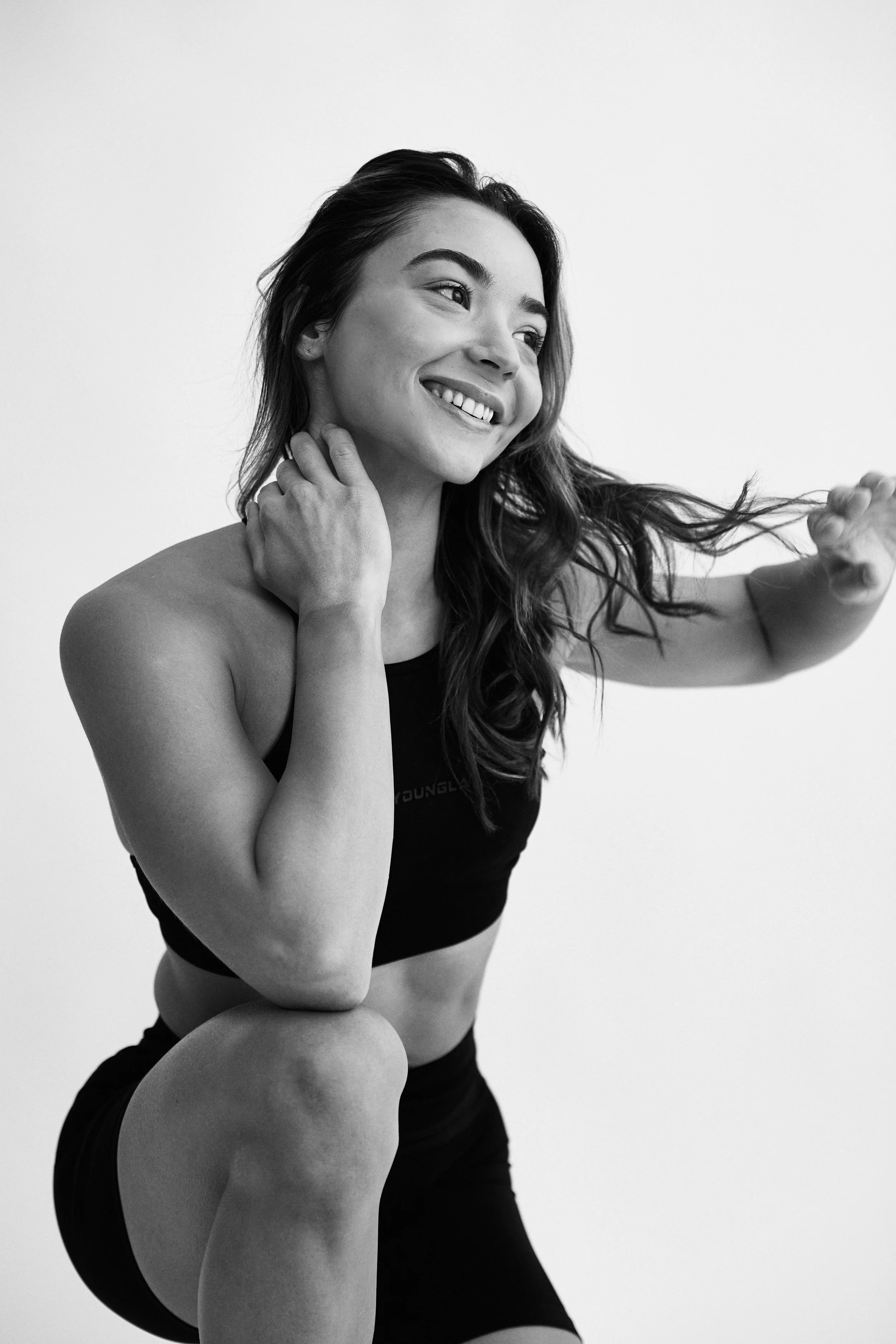 Black and white portrait of a woman smiling, with long wavy hair, wearing a sports bra and shorts, touching her neck with one hand and holding her hair with the other.