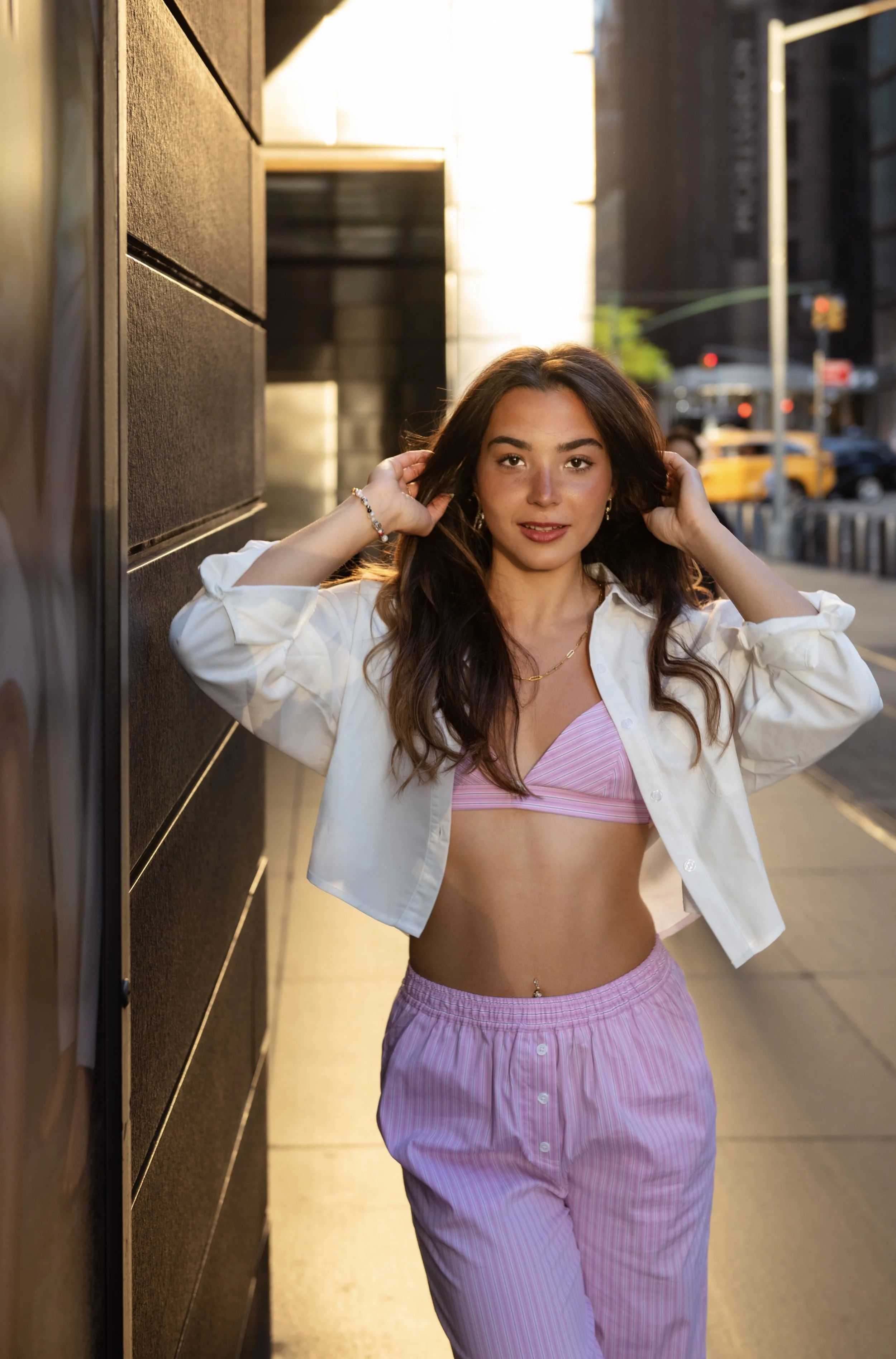 A young woman with long dark hair and light skin posing outdoors on a city sidewalk during sunset, wearing a pink striped crop top, pink striped pants, a white jacket, earrings, and a bracelet.