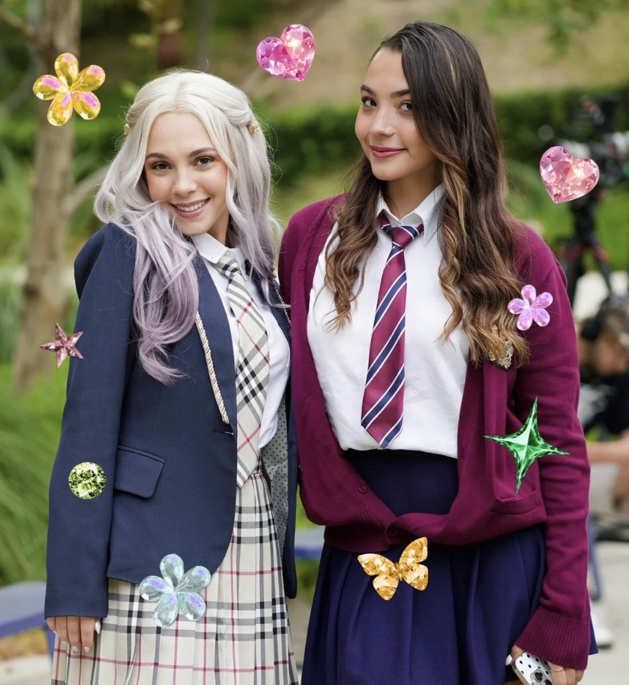 Two teenage girls in school uniforms standing outdoors, smiling. One has long, wavy dark brown hair, wearing a maroon blazer and striped tie. The other has long, wavy light-colored hair with lavender tips, wearing a navy blazer and plaid skirt. Decor