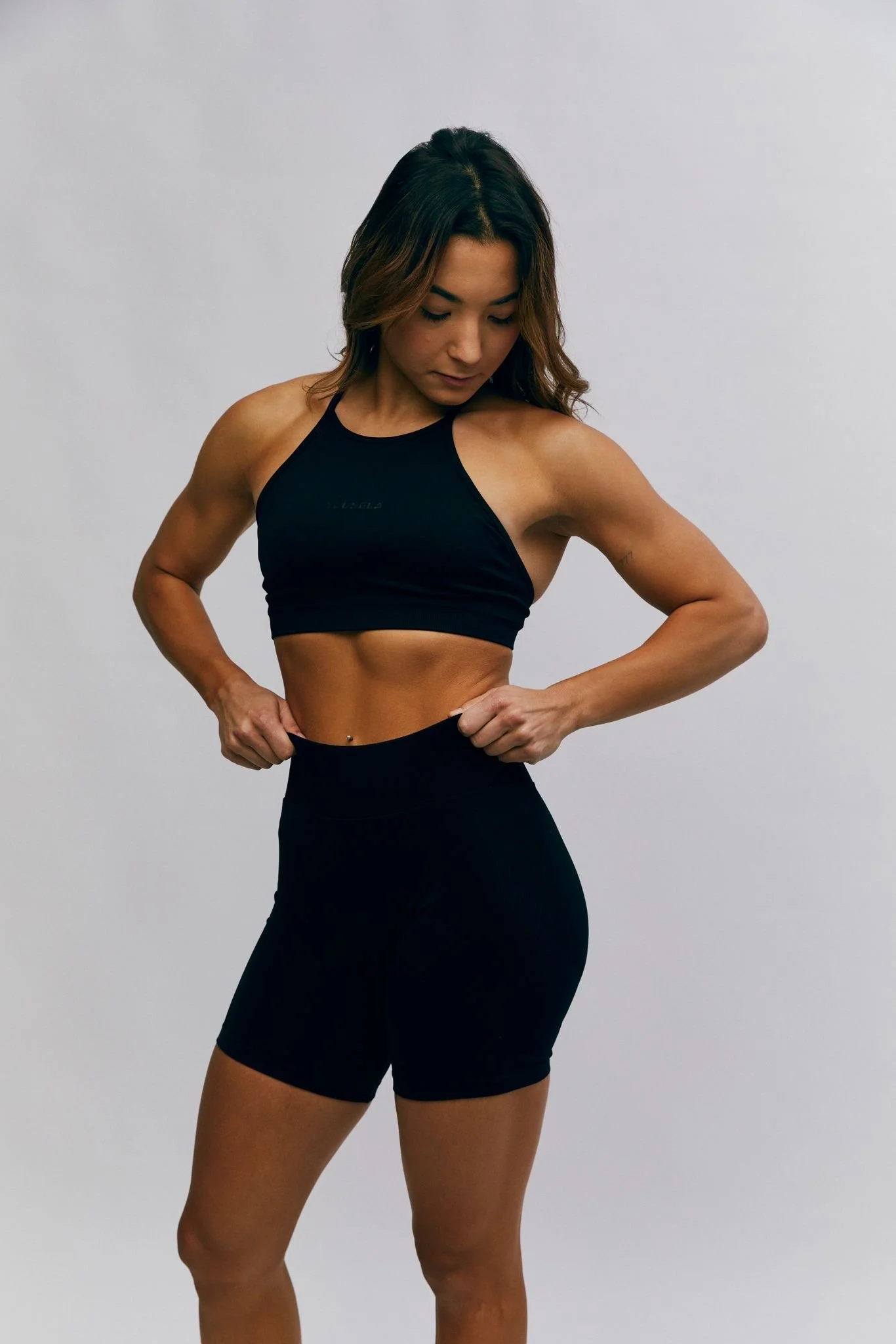 A woman in a black sports top and shorts is standing against a plain gray wall, looking down and to her right, with her hands on her hips.