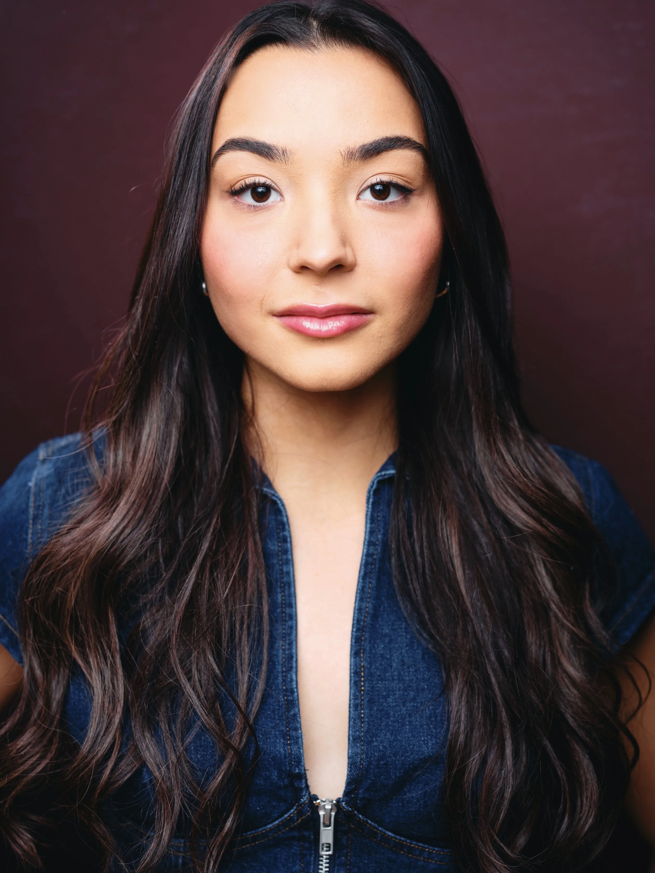 Close-up photo of a young woman with long dark hair, wearing a denim jacket, standing against a dark maroon background.