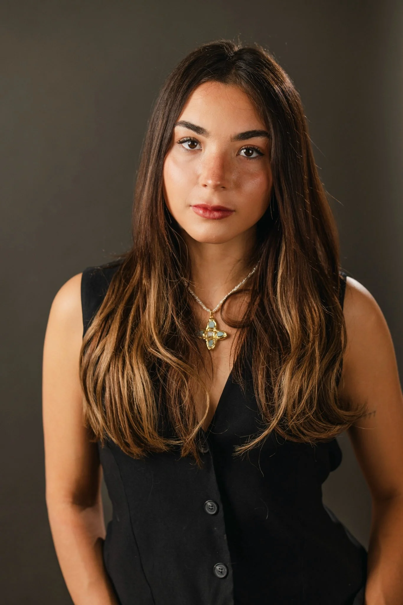 A young woman with long, wavy brown hair wearing a black sleeveless dress with buttons and a necklace with a cross pendant, standing against a dark gray background.
