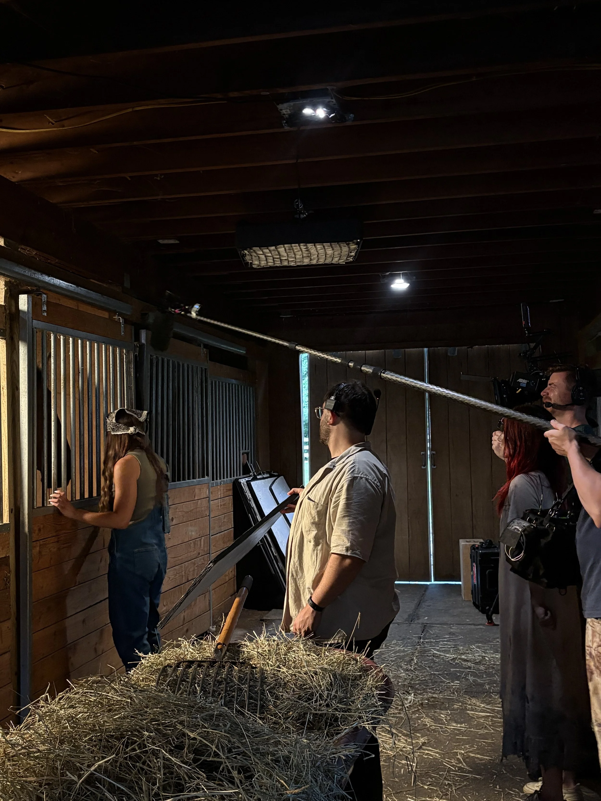 People on a film or television set, standing near a wooden stable with hay, using cameras and microphones.
