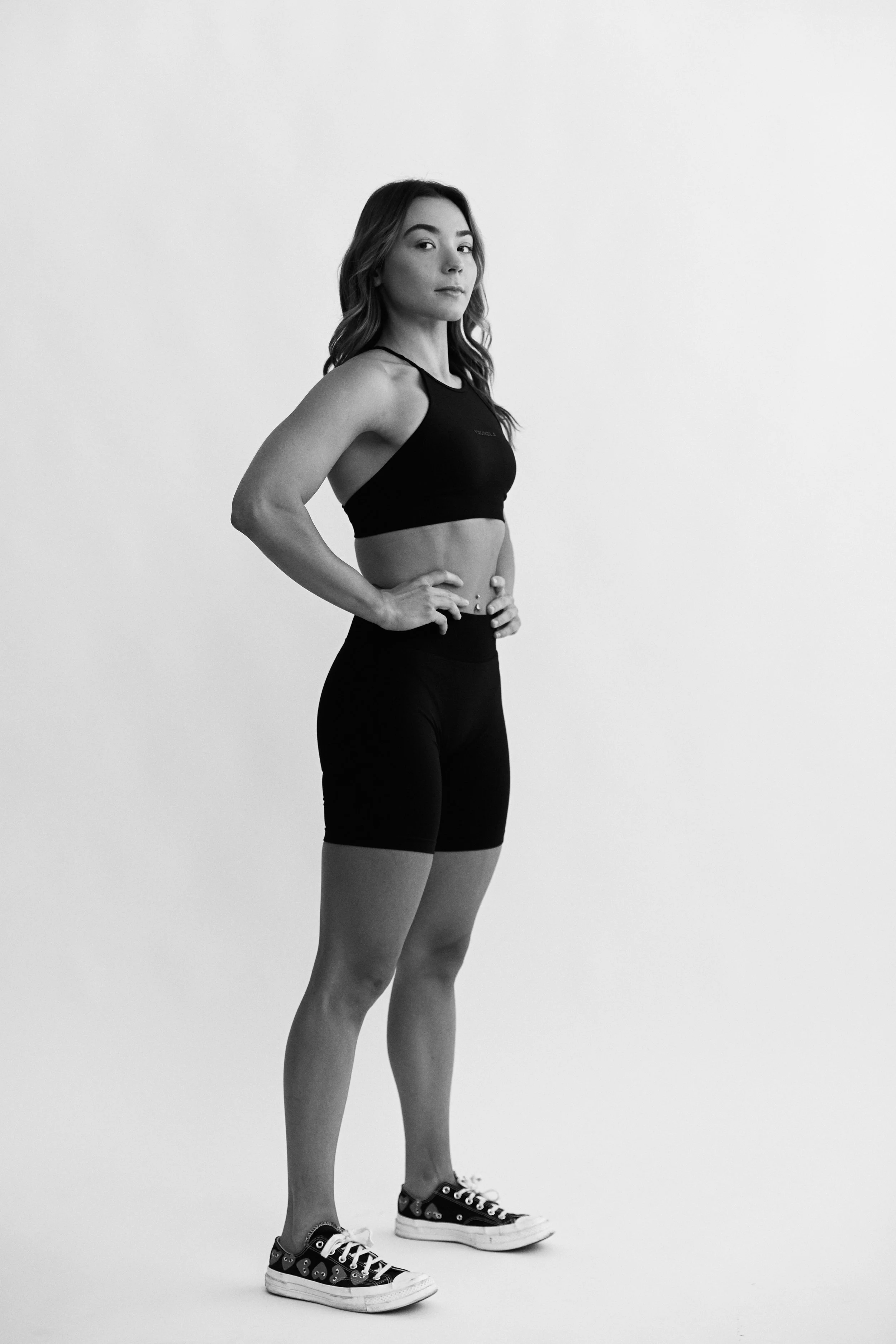 A black and white photograph of a young woman in athletic attire standing in a studio with a plain background. She is wearing a sports bra, high-waisted shorts, and patterned sneakers. She has long, wavy hair and gazes confidently into the camera wit
