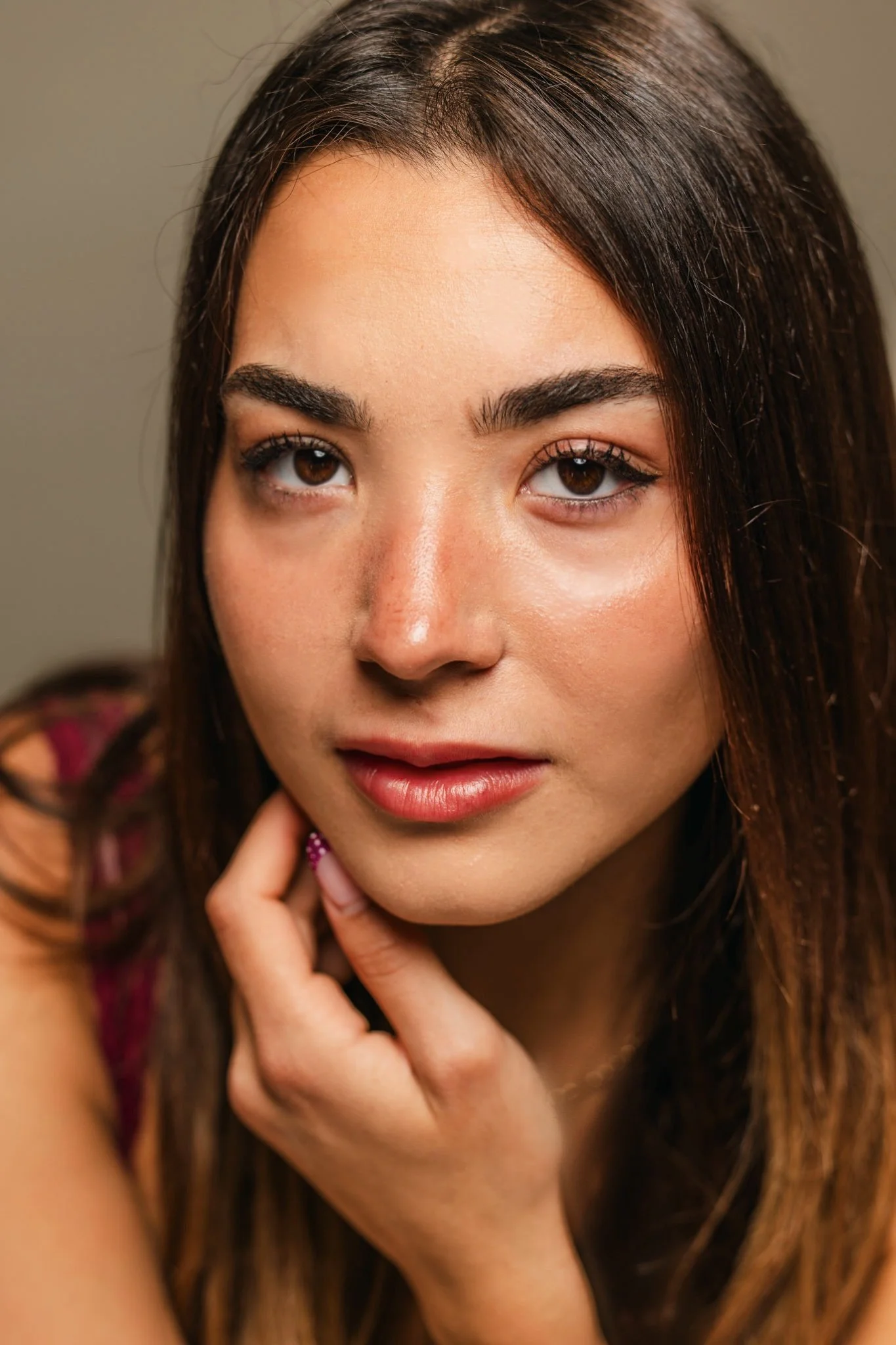 A close-up portrait of a woman with long brown hair, wearing makeup, and posing with her hand near her face.