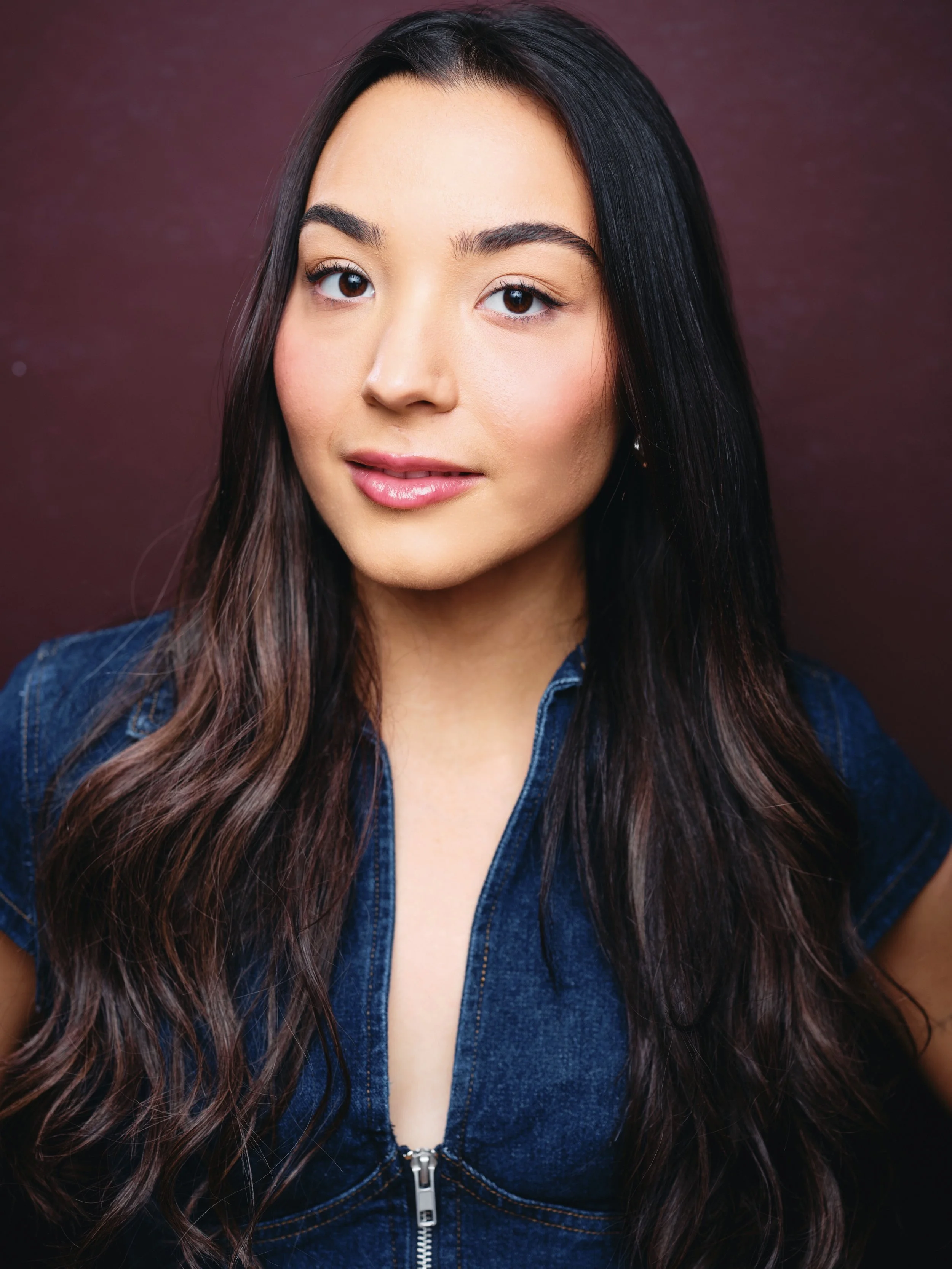 Close-up of a young woman with long, dark, wavy hair wearing a denim jacket with a zipper, against a purple background.
