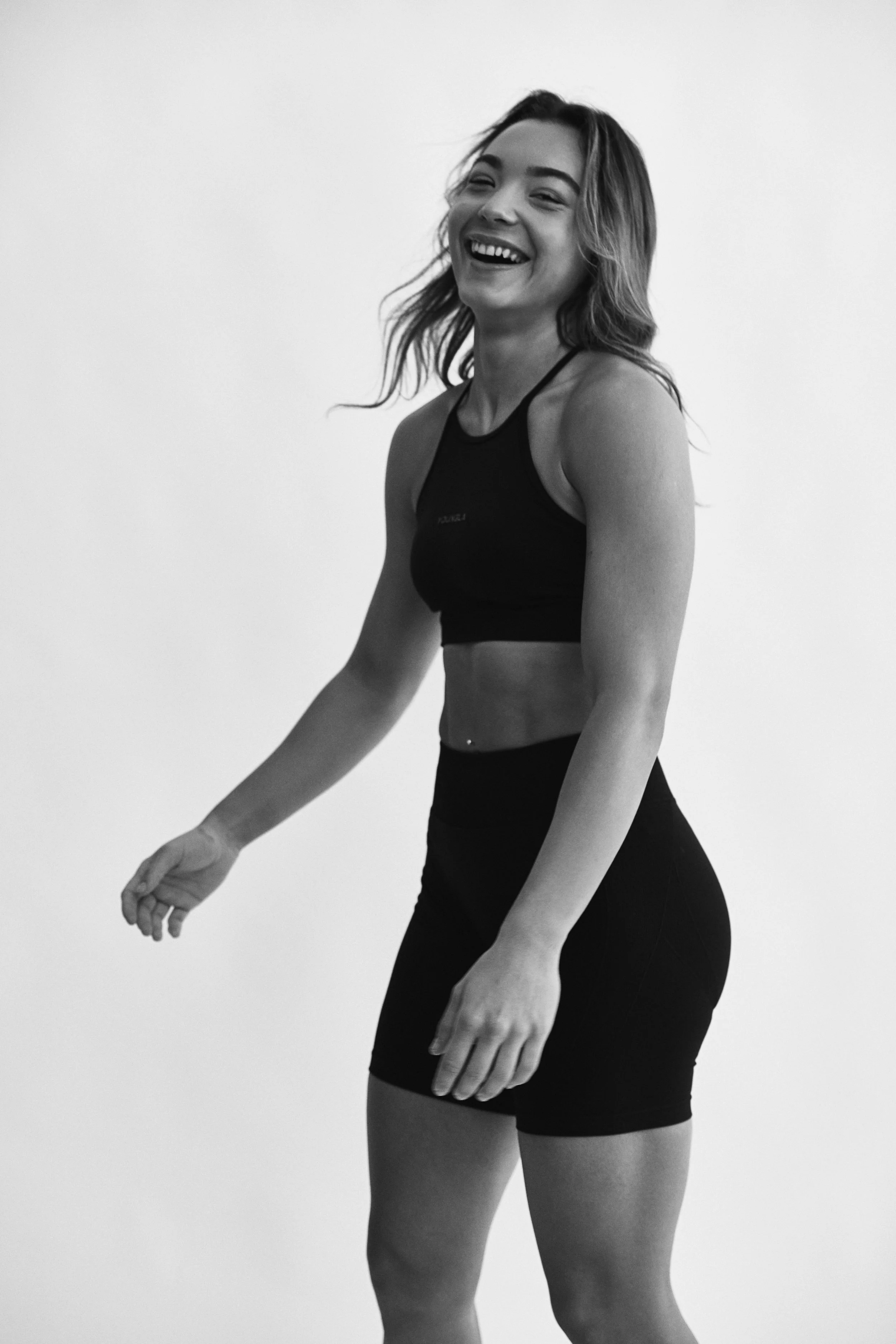 A woman smiling and wearing workout clothes, including a sports bra and leggings, standing against a plain white background.