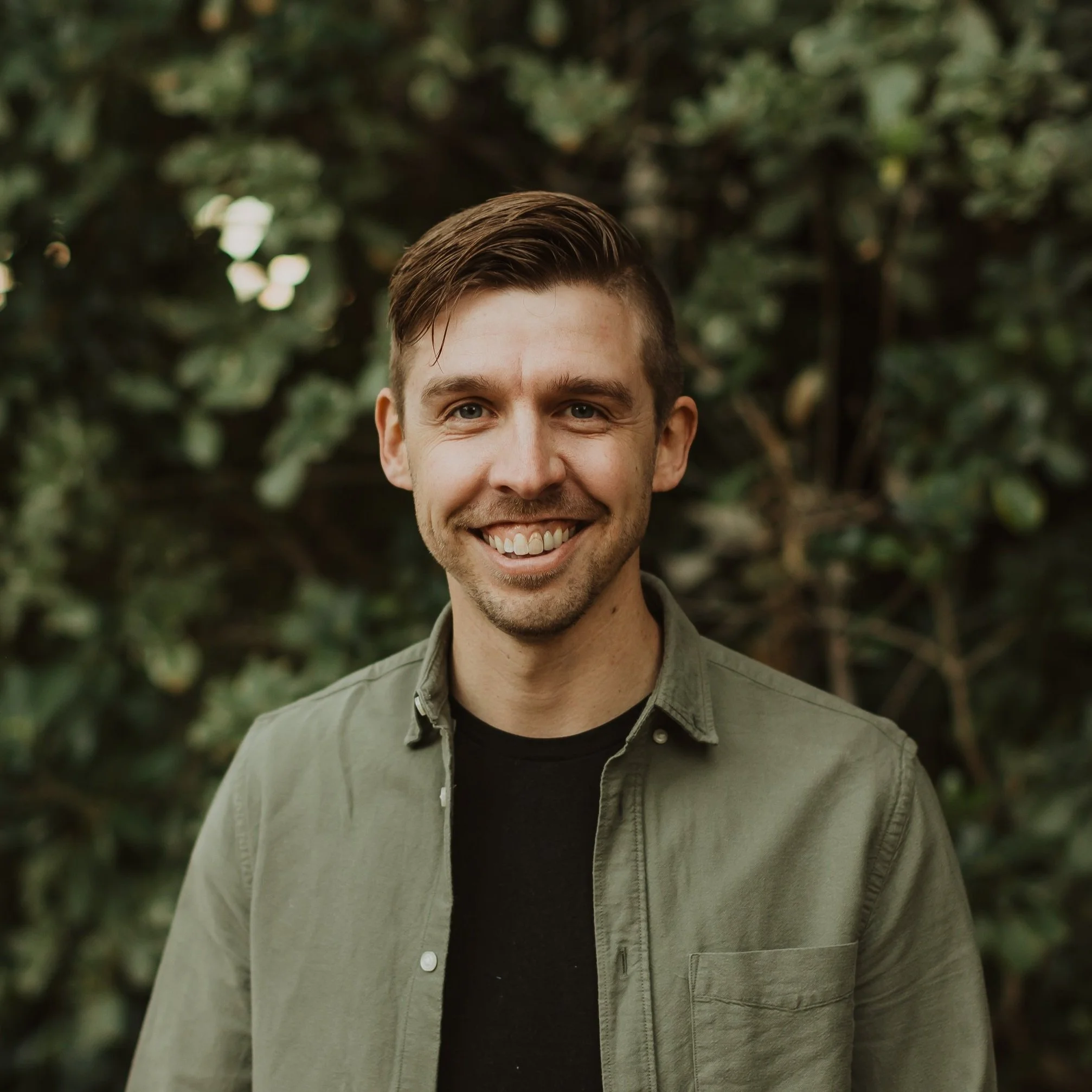 A smiling young man with short brown hair, wearing a green button-up shirt over a black t-shirt, standing outdoors in front of greenery.