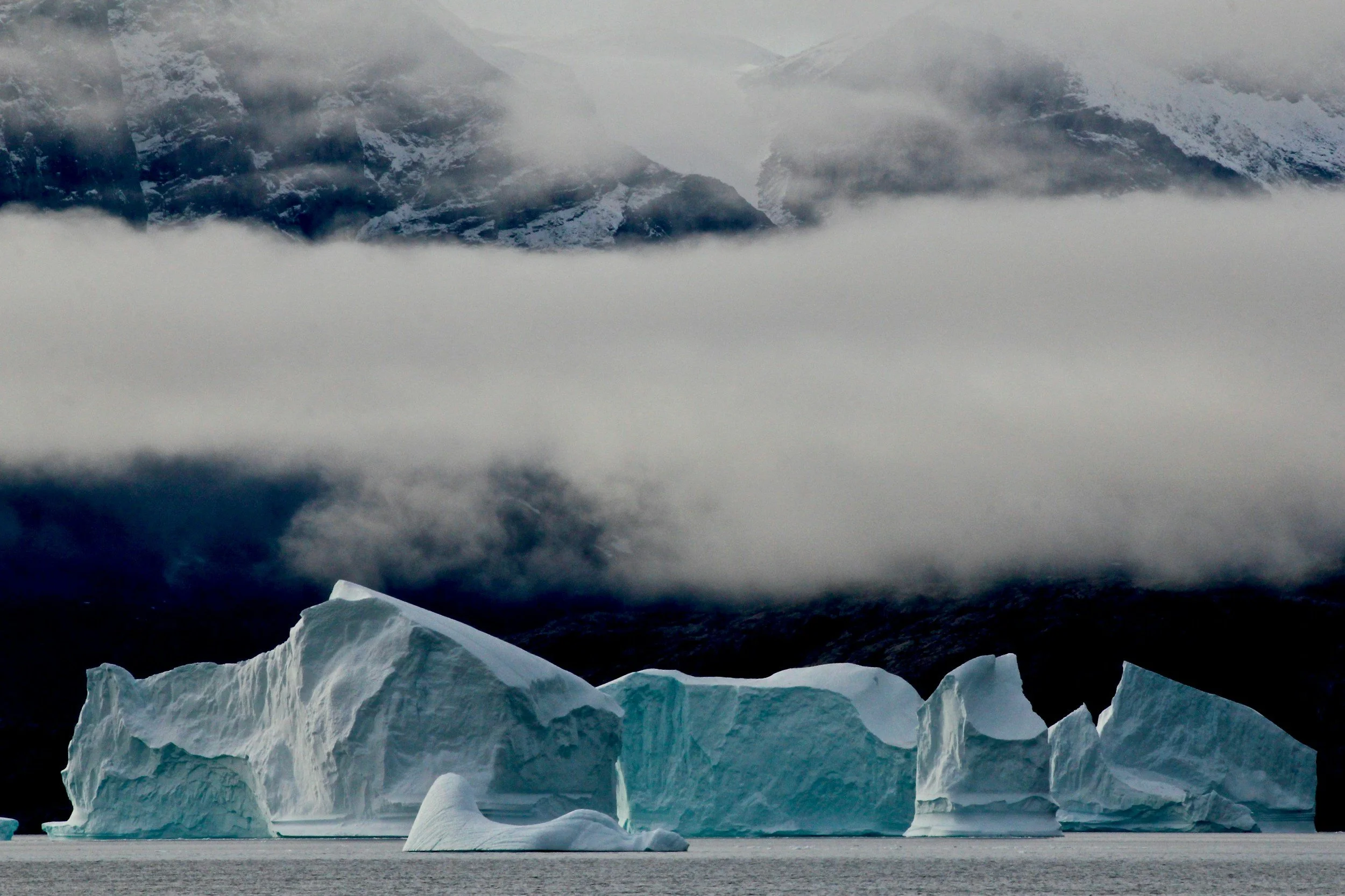 Icebergs floating in a dark ocean with snow-capped mountains and cloudy sky in the background.
