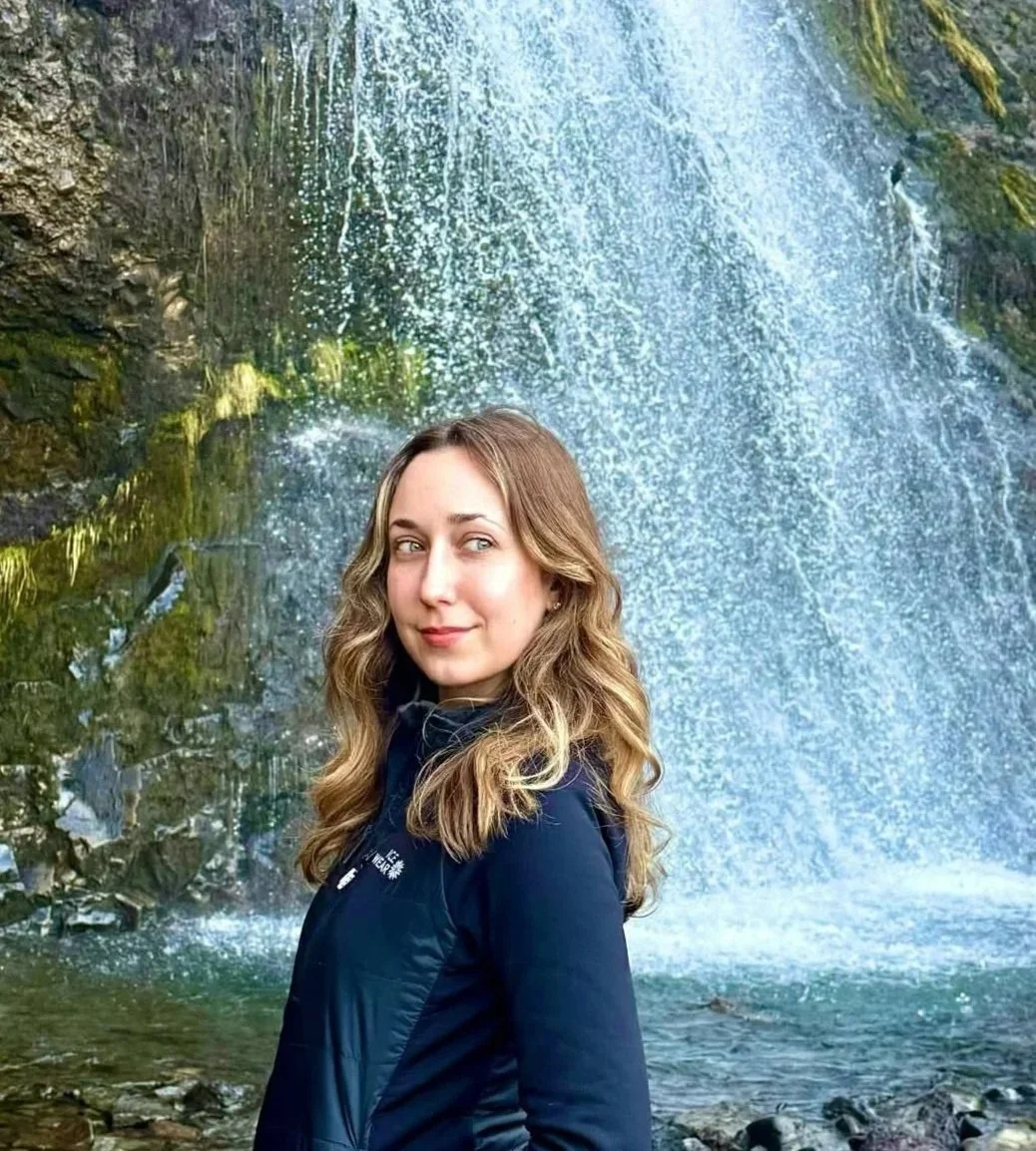 A young woman with wavy blonde hair smiling at the camera, standing in front of a waterfall surrounded by rocks and moss.