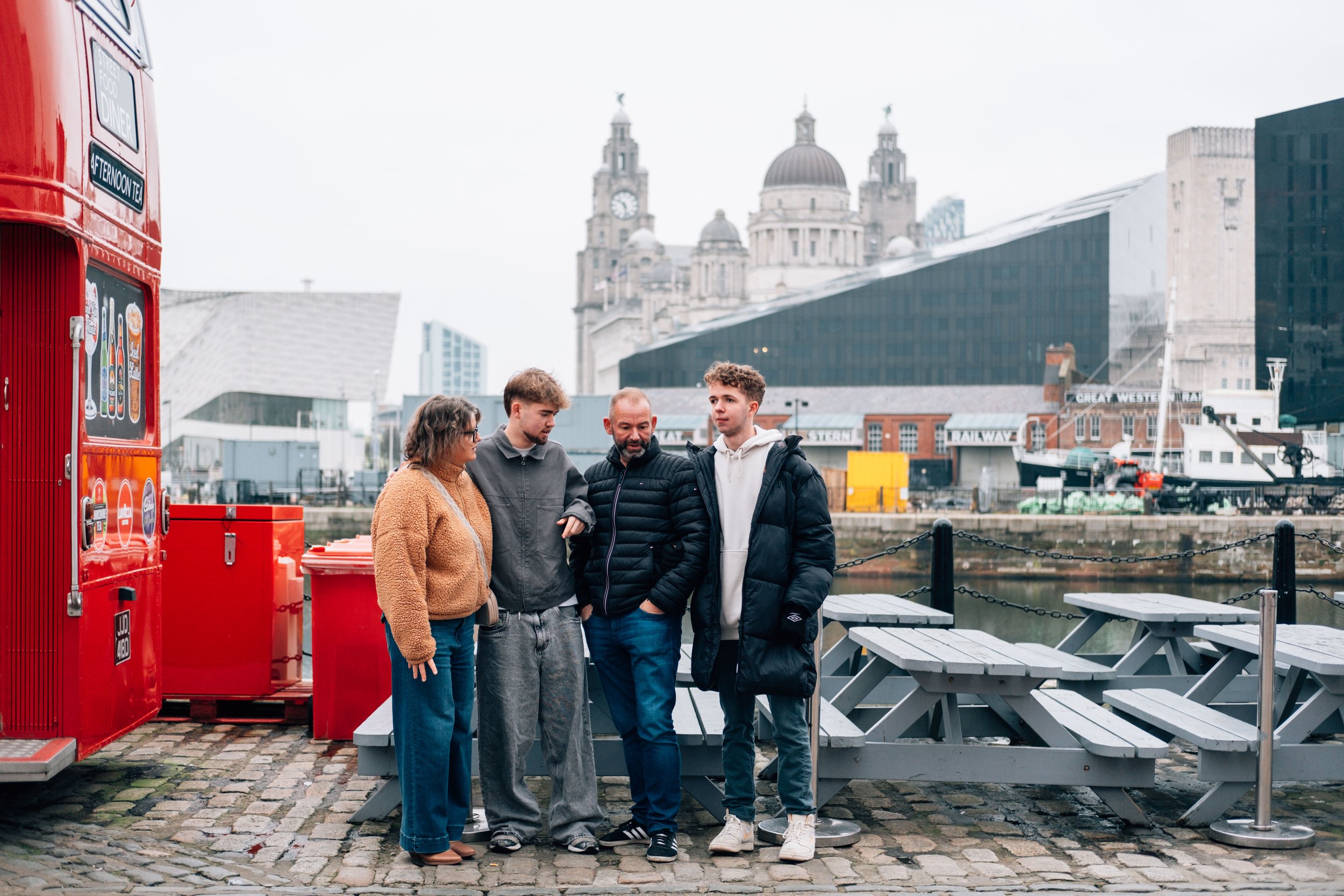 Four people standing on a cobblestone path near water, with a red food bus on the left and historic buildings in the background, including a church with domes and towers.