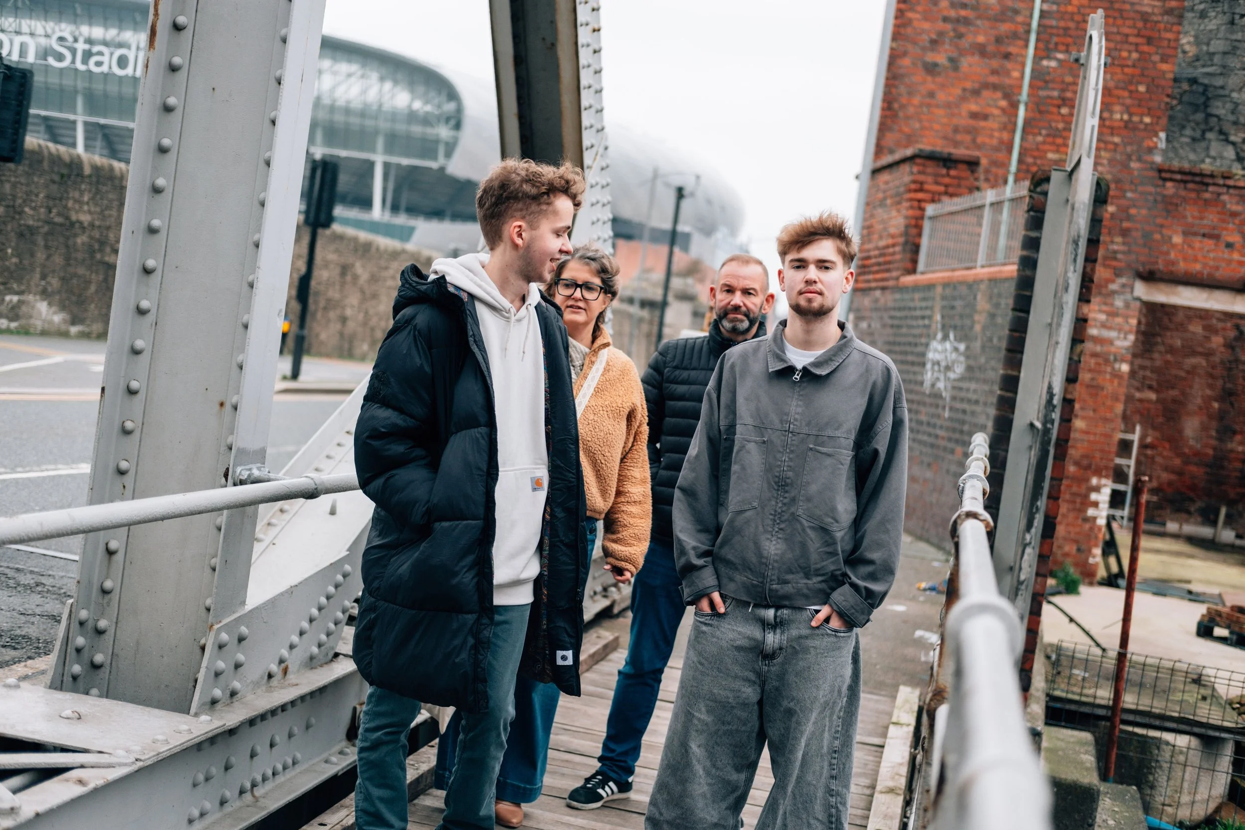 Four people standing on a metal bridge outdoors, with buildings in the background.