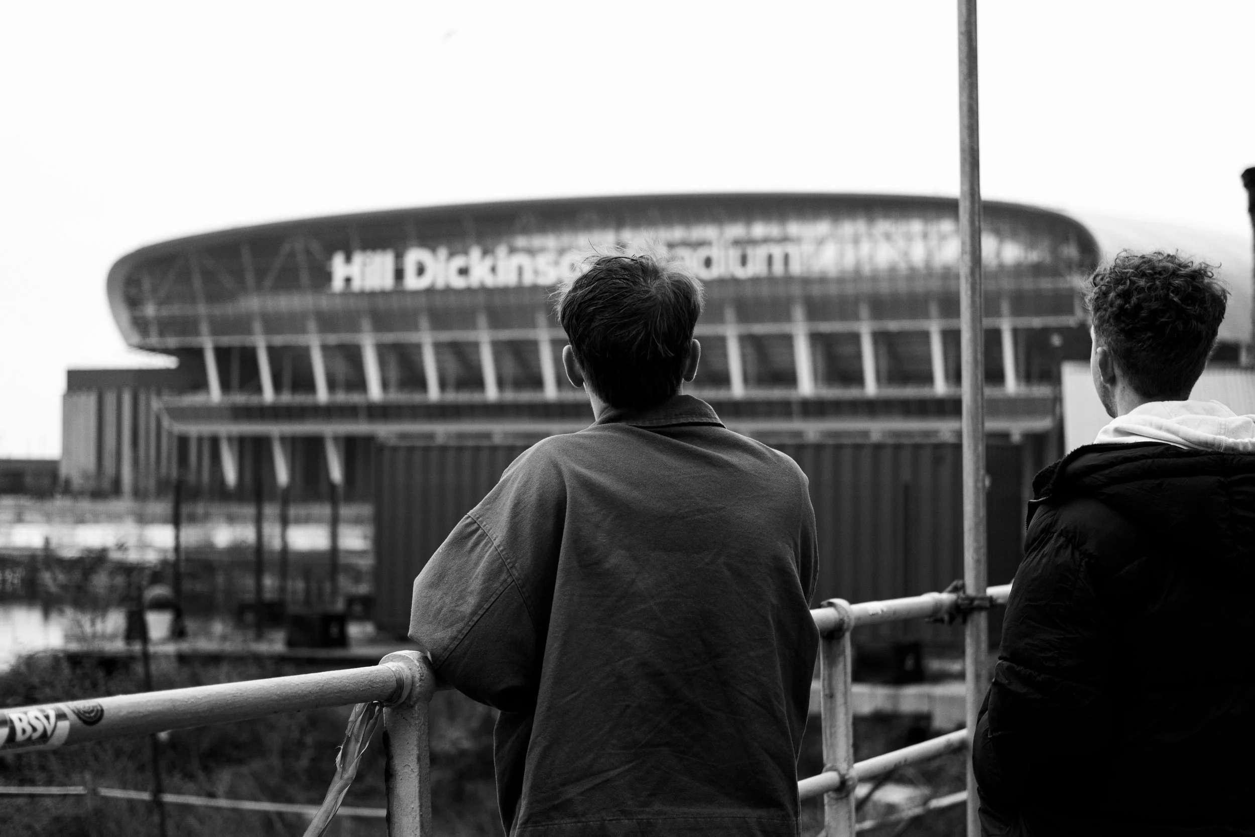 Two men are standing by a railing, looking at the Hillsborough Stadium in Sheffield, England.