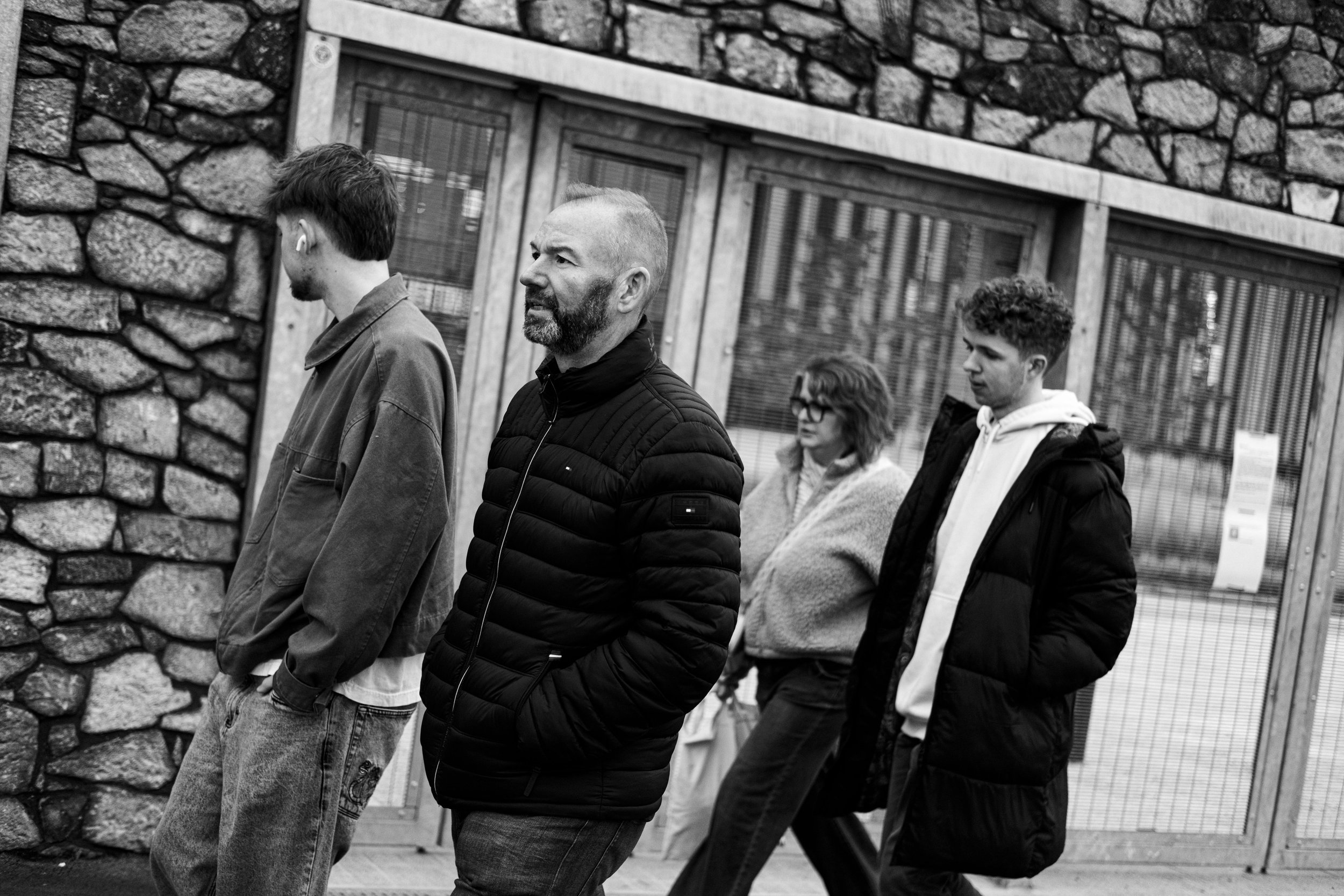 Four people walking on sidewalk in front of a stone building with a glass door, with three men and one woman, all casually dressed.