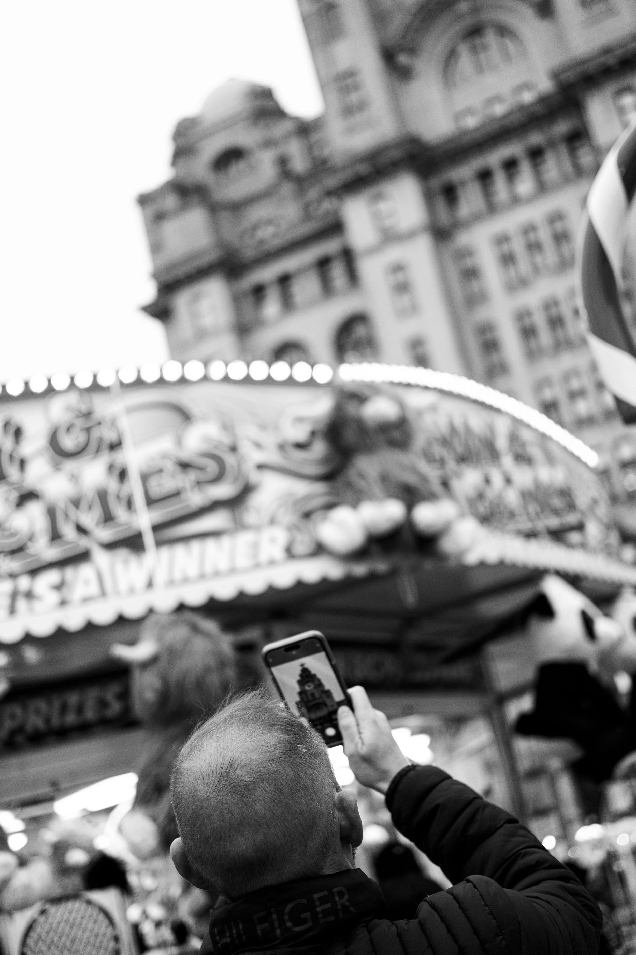 A person with short hair taking a photo of a carousel with their smartphone at a fair, with a historic building in the background in black and white.