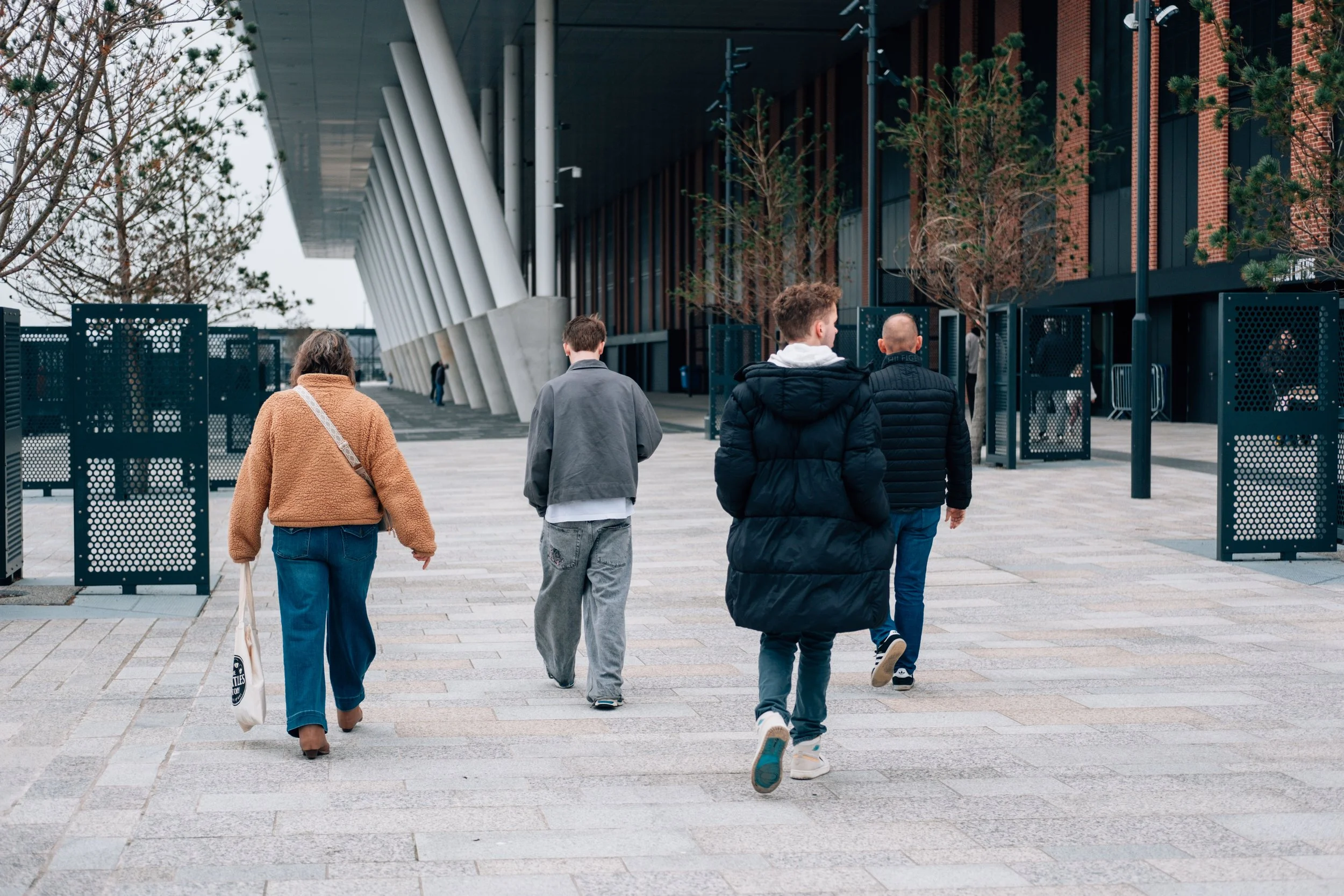 A group of five people walking away from the camera on a city sidewalk near a modern building with large white structural columns and leafless trees.