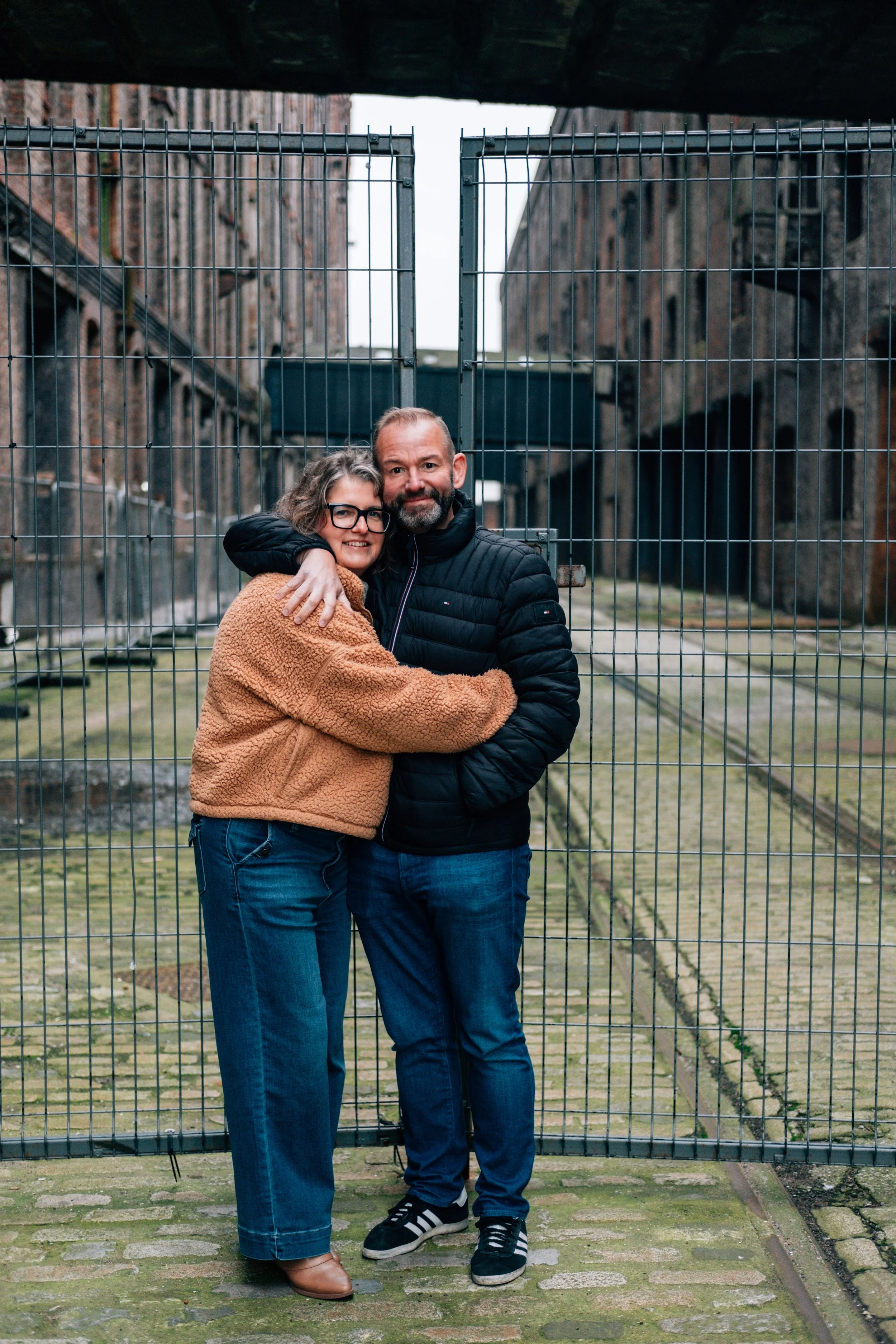 A couple embracing in front of a metal gate in an outdoor urban area with brick buildings.