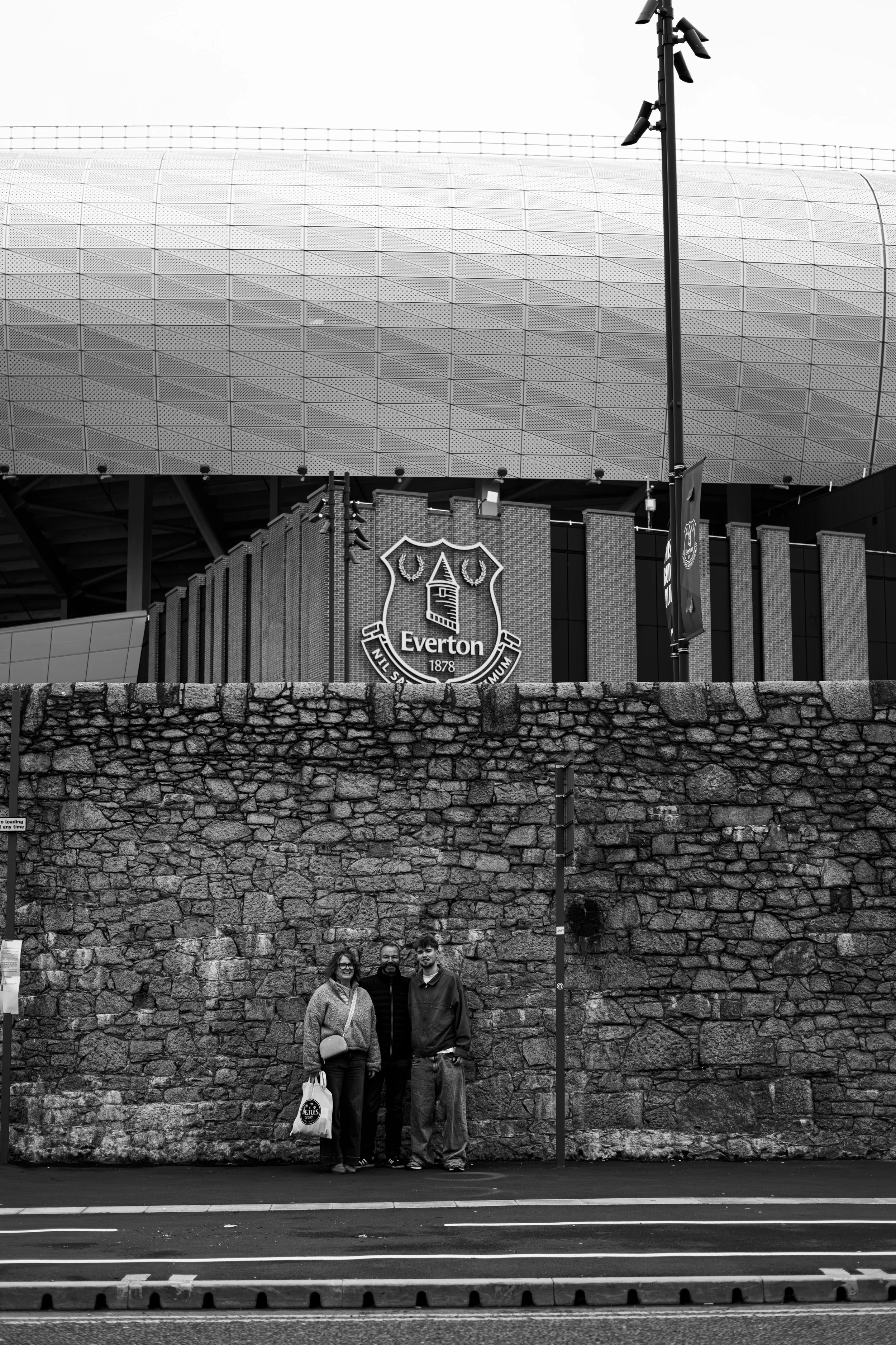Black and white photo of three people standing against a stone wall outside Everton football stadium, with the stadium's modern design and the club's crest visible in the background.