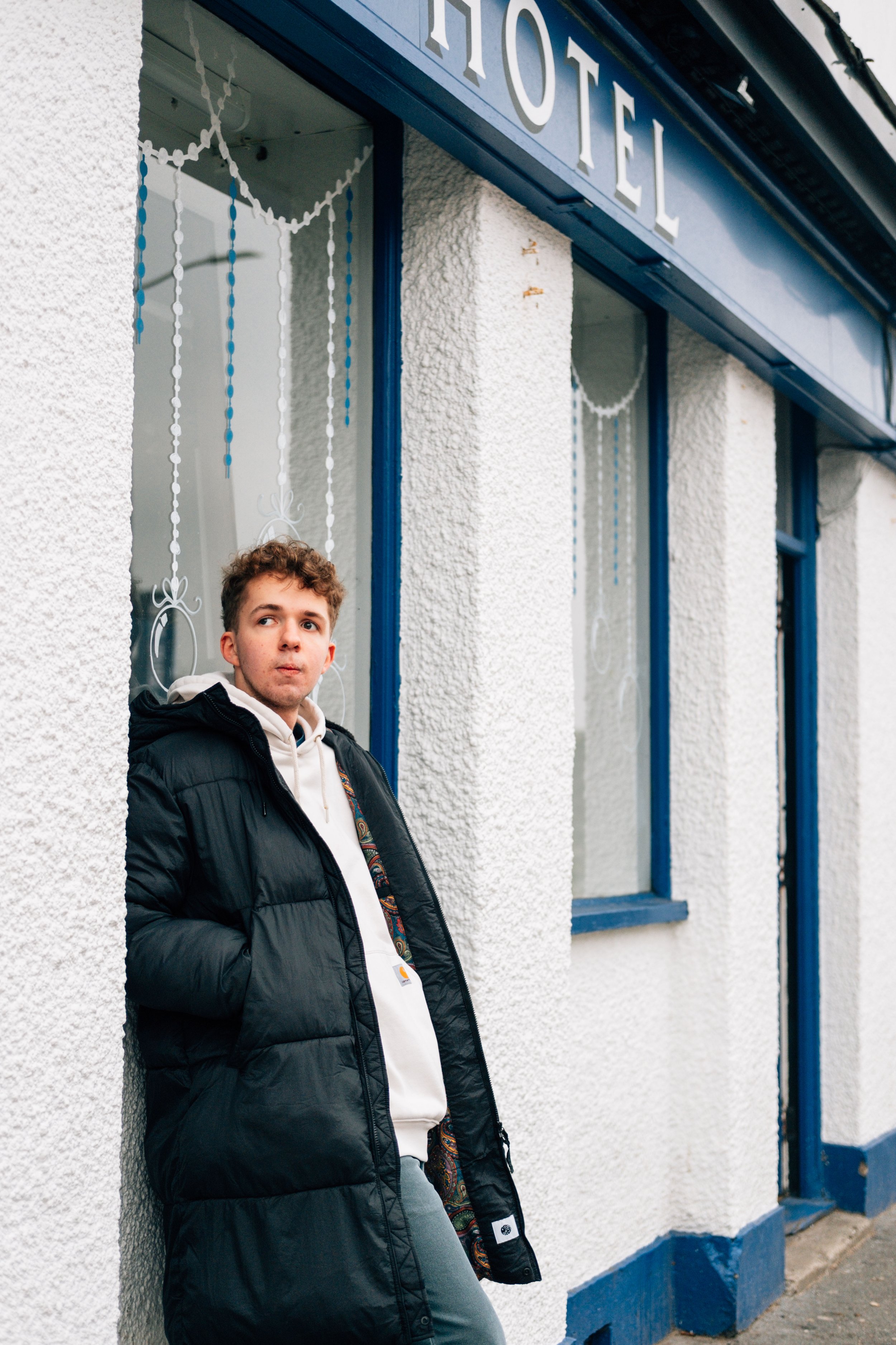 A young man standing outside a white textured building with a sign that says 'HOTEL.' He is wearing a black puffer jacket and a white hoodie, with hands in his pockets, looking off to the side.
