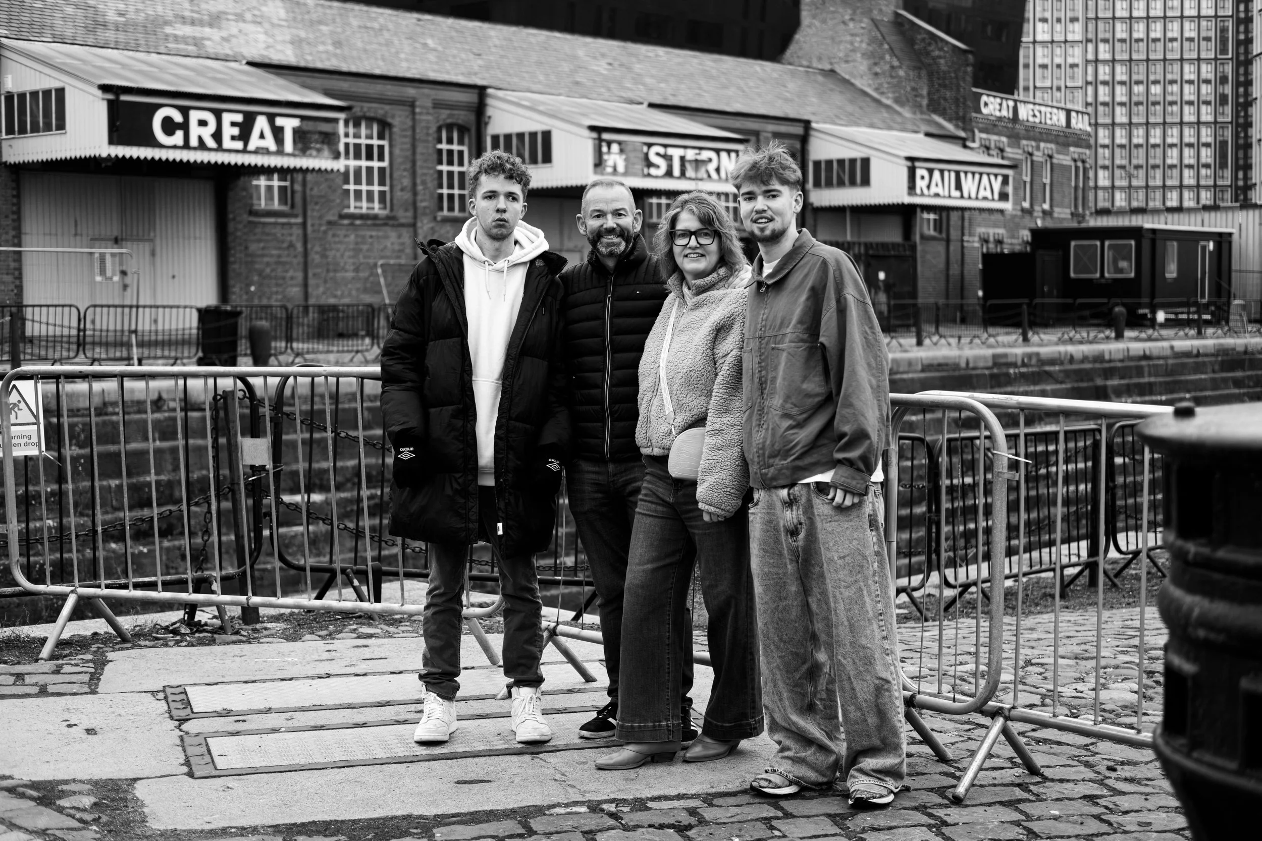 Four people stand near a canal with barriers, behind old brick buildings and a sign that reads 'Great Western Rail' in a city setting.