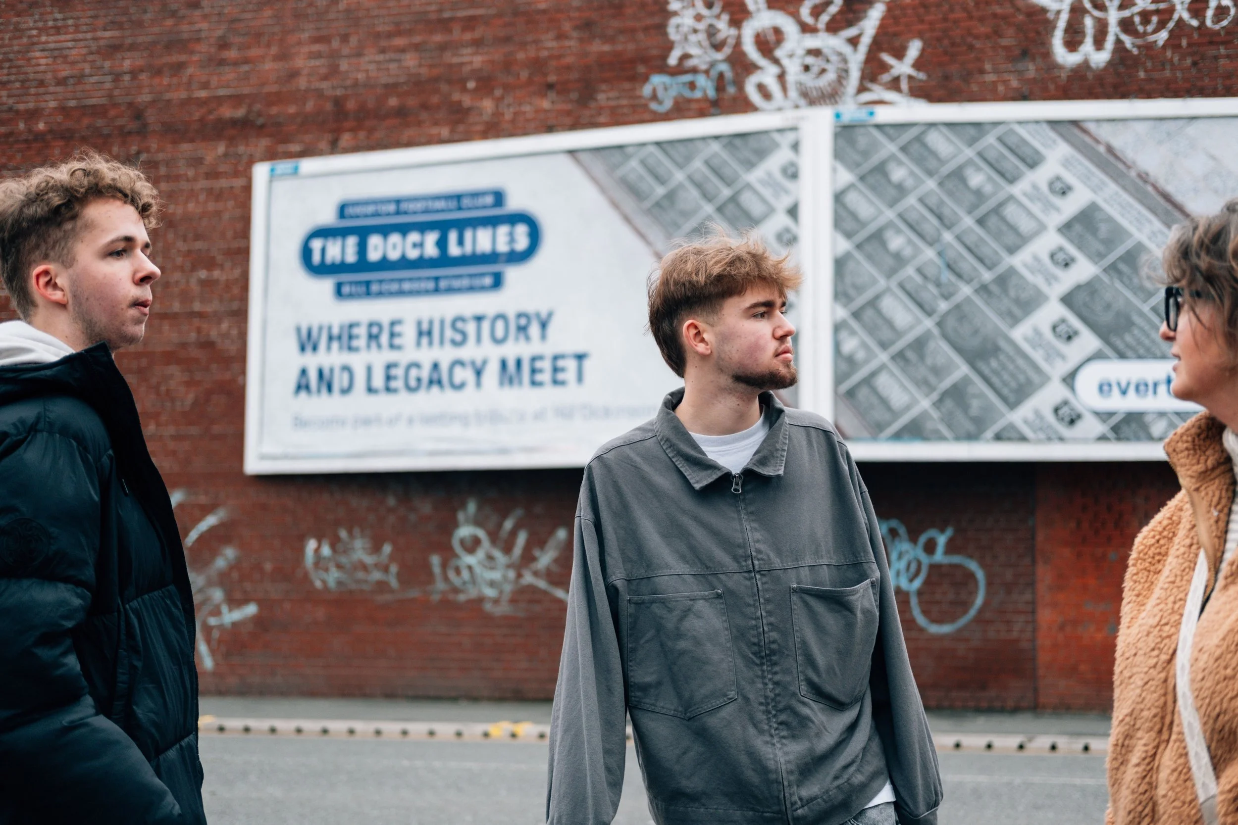 Three young adults standing on the street near a brick wall with graffiti and a large sign that reads 'The Dock Lines: Where History and Legacy Meet'.