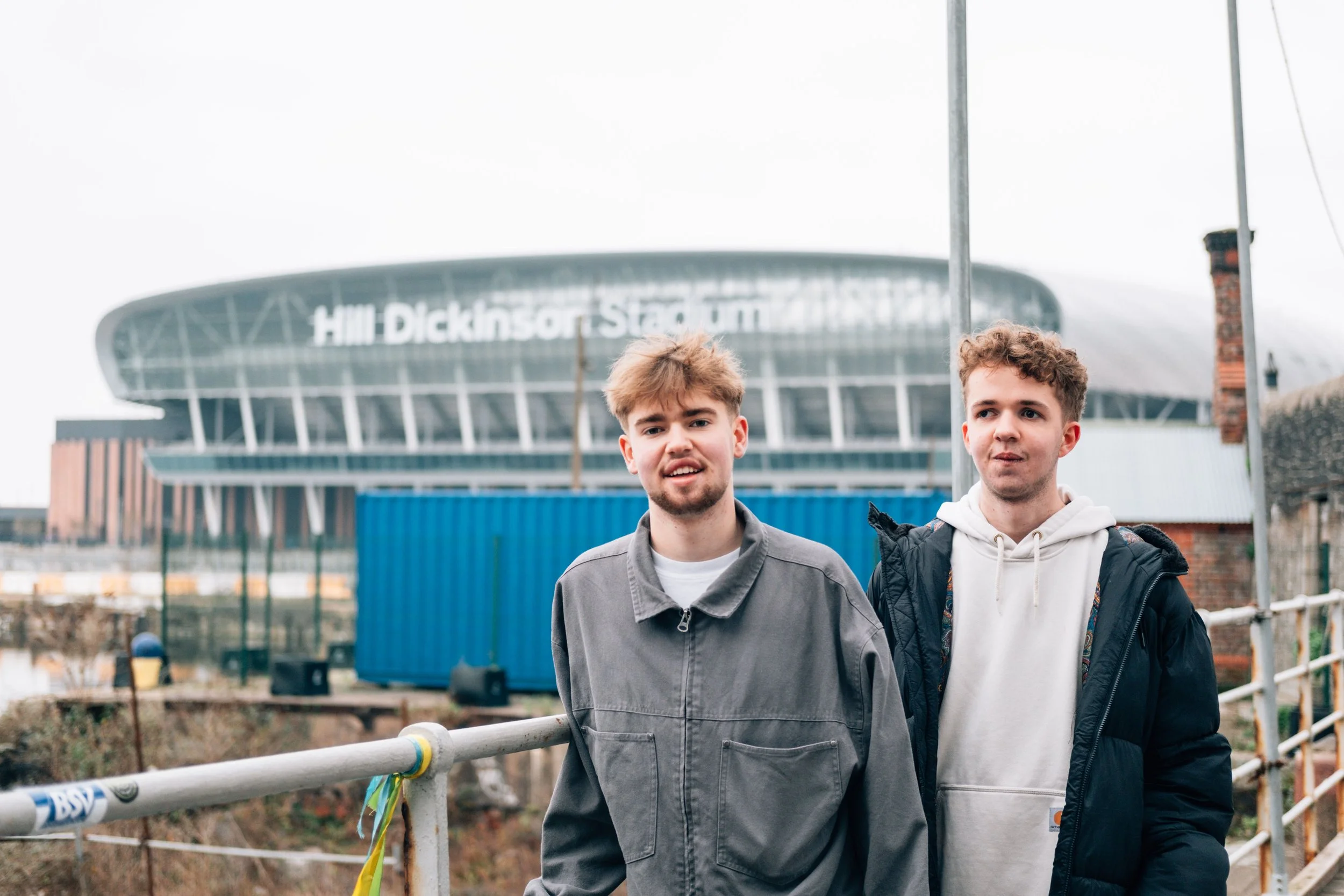 Two young men standing outdoors with a stadium in the background that reads 'Hill Dickinson Stadium'.