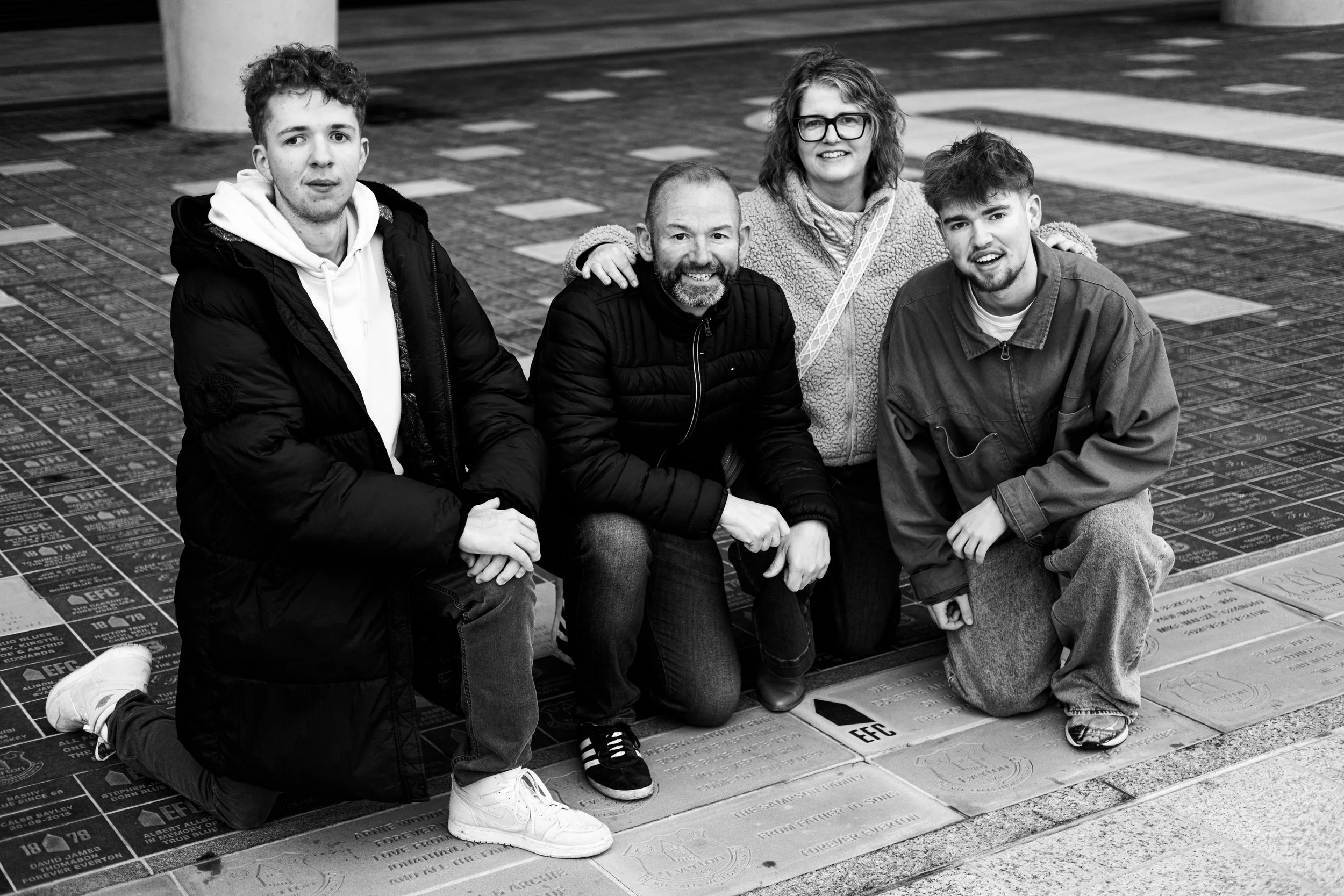 Four people kneeling and smiling on a memorial sidewalk, with engraved plaques and markings.