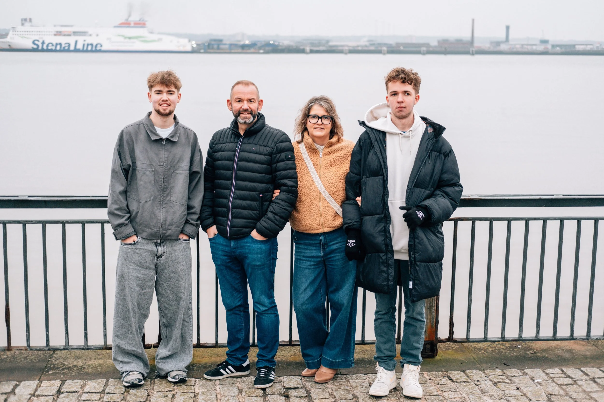 Four people standing outdoors on a dock near water, with a ferry and industrial buildings in the background.