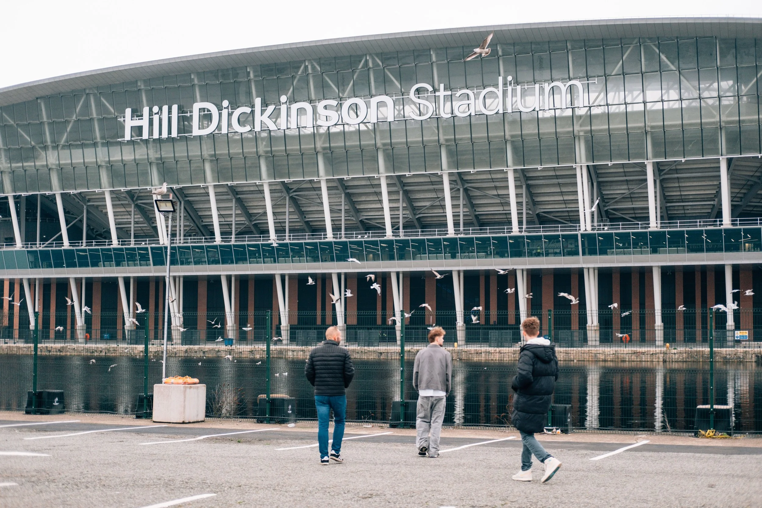 People walking outside Hill Dickinson Stadium, a large modern building with a glass exterior and a sign displaying its name.