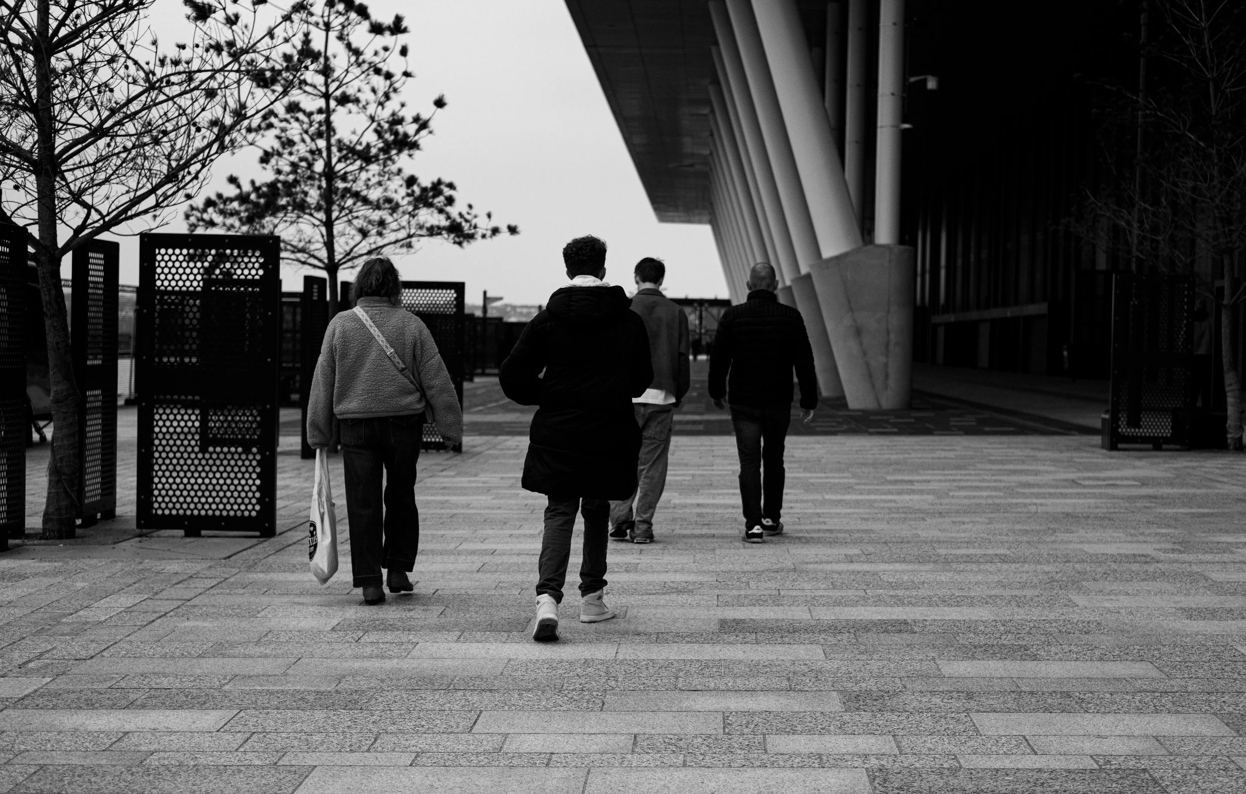A black-and-white photo of five people walking outdoors near a modern building with angled columns, trees, and a pathway.