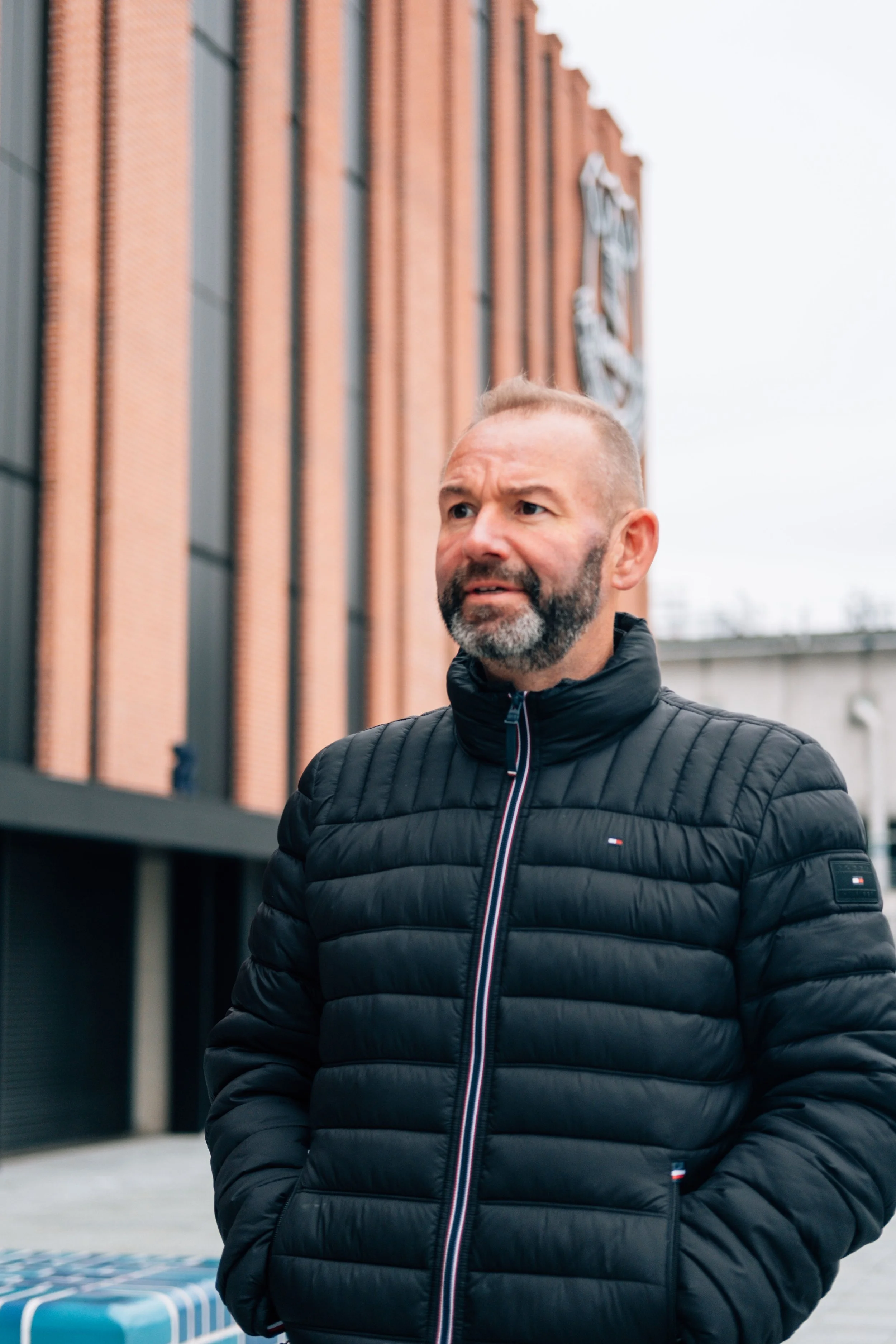 A middle-aged man with a beard and short hair wearing a black quilted jacket standing outdoors in front of a modern brick building.