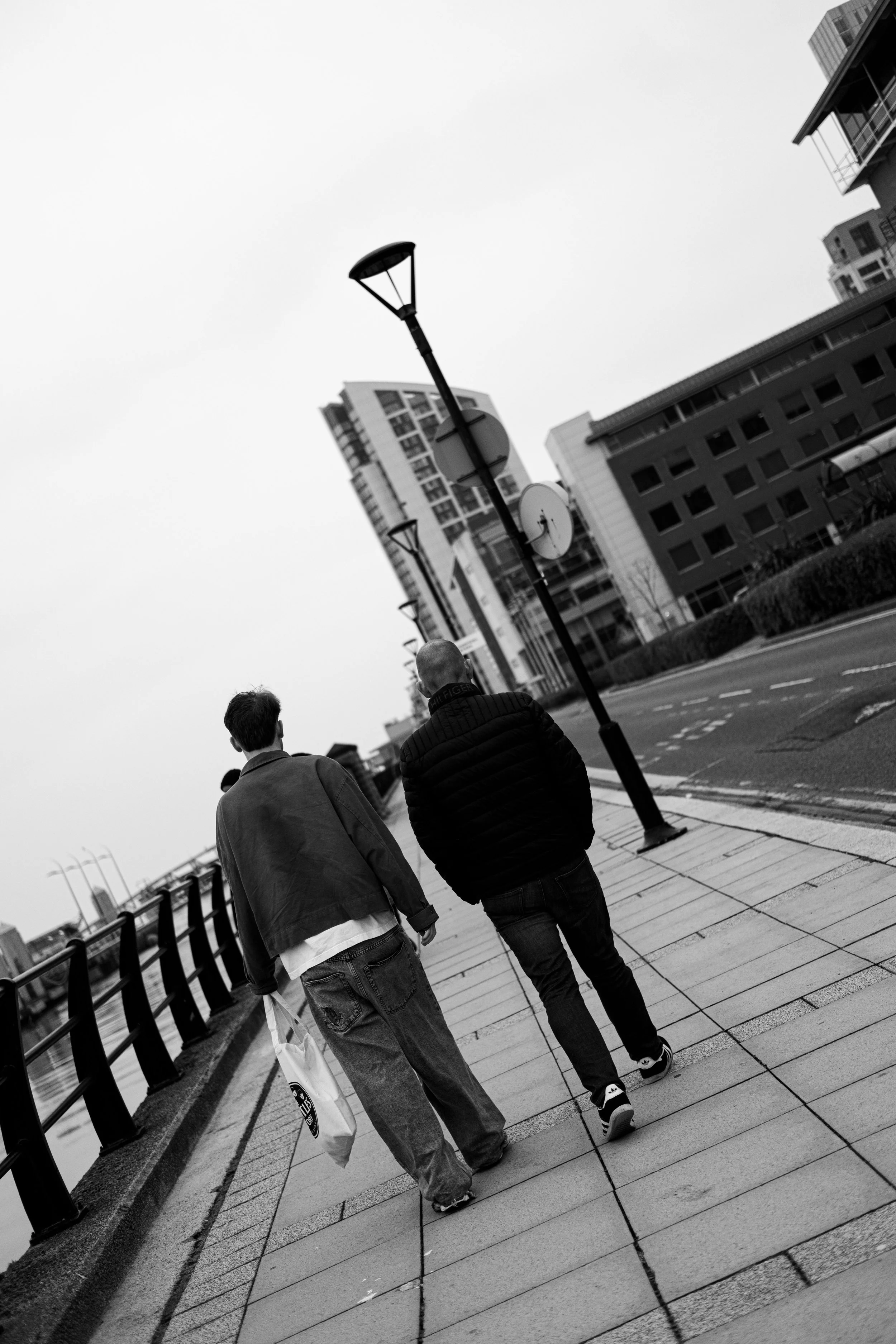 Two people walk along a city sidewalk beside a river, with modern buildings and street lamps. The scene is in black and white.