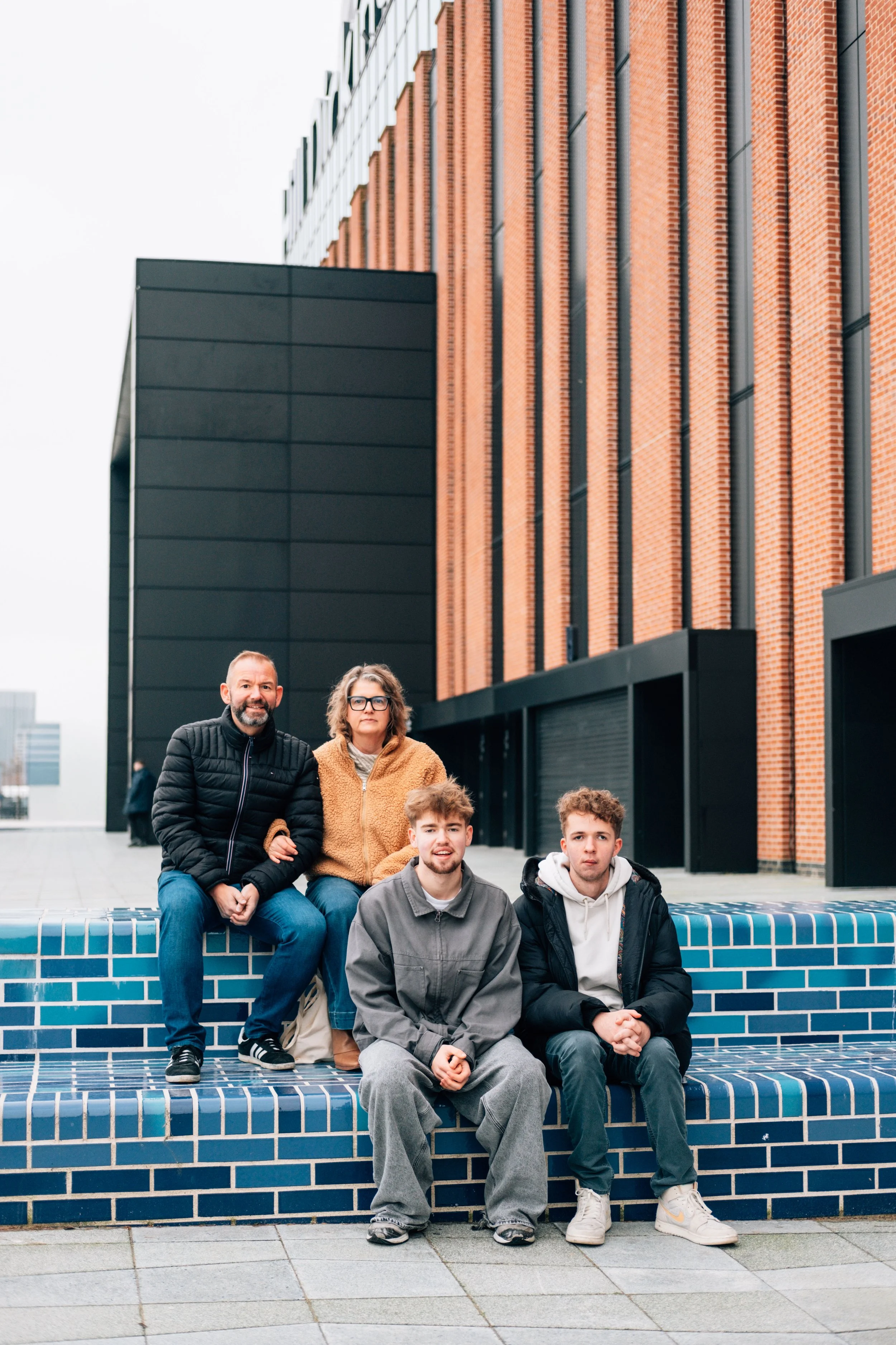 Group of five people sitting on blue tiled bench in front of modern building with brick and black exterior walls.