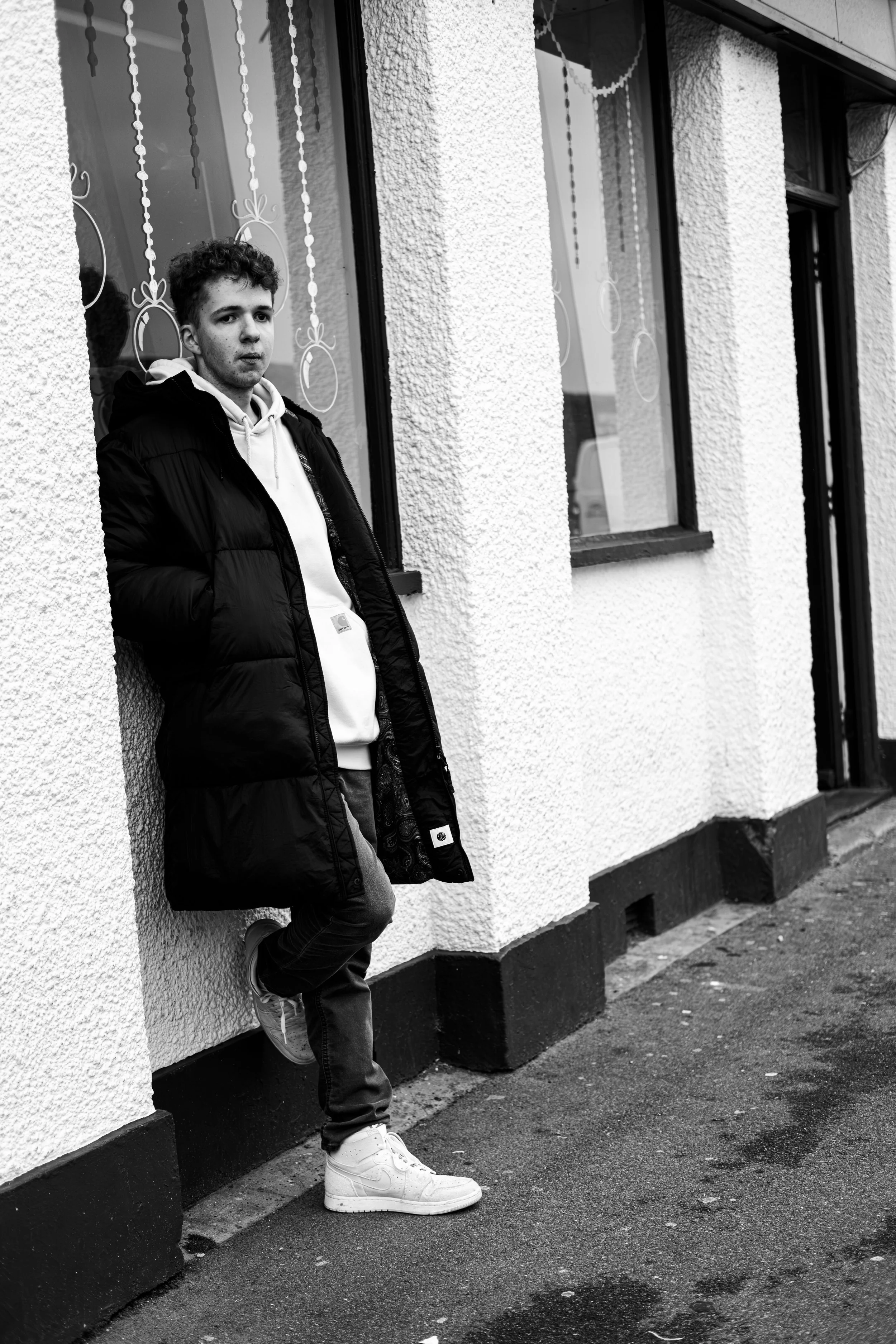 A young man is standing outside on the sidewalk, leaning against the textured wall of a building. He is wearing a puffer jacket, hoodie, jeans, and sneakers, with his hands in his pockets and looking slightly to the side.