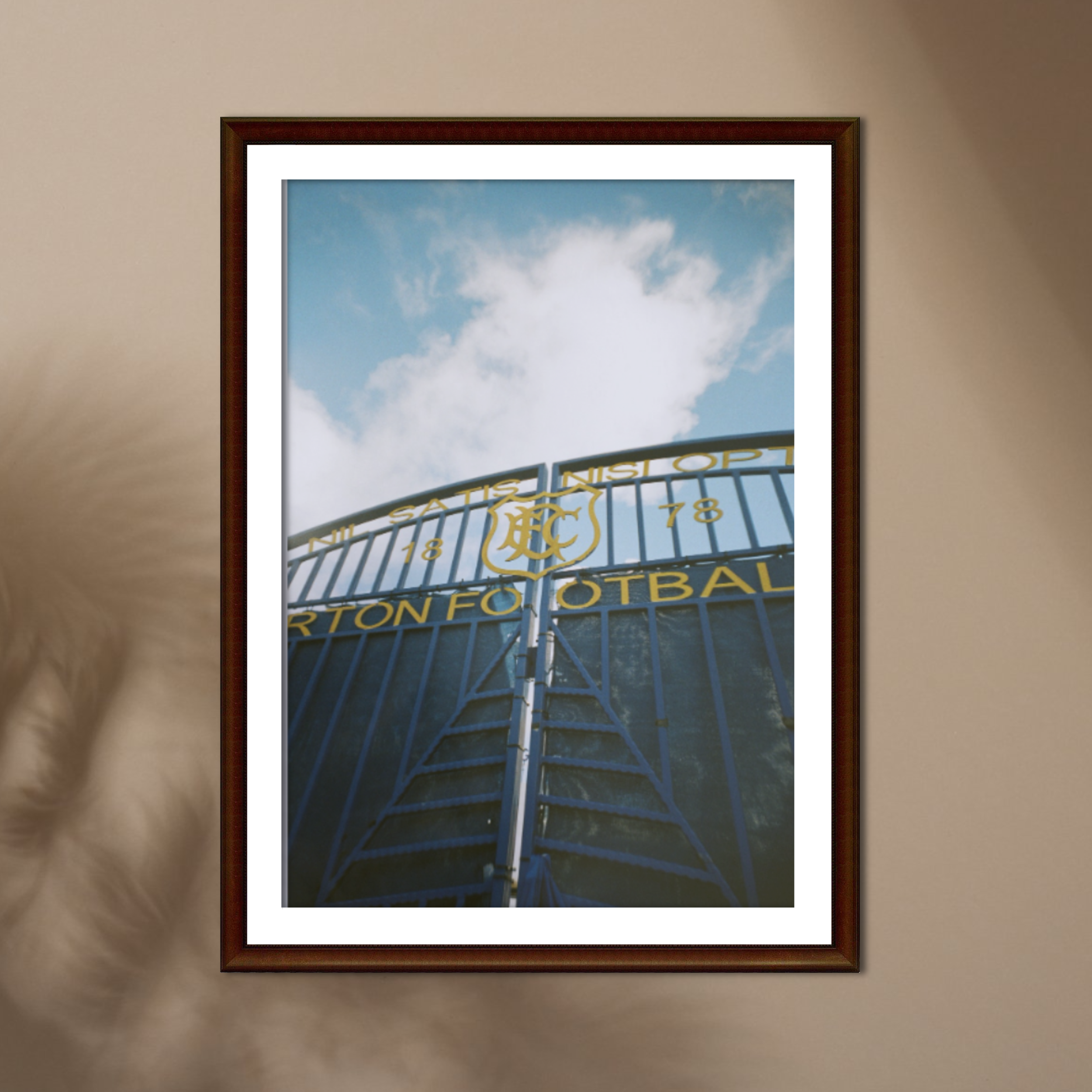 Photograph of a stadium entrance gate with blue metal fencing, featuring a logo, date 1878, and part of a phrase in yellow lettering, with a blue sky and white clouds overhead.