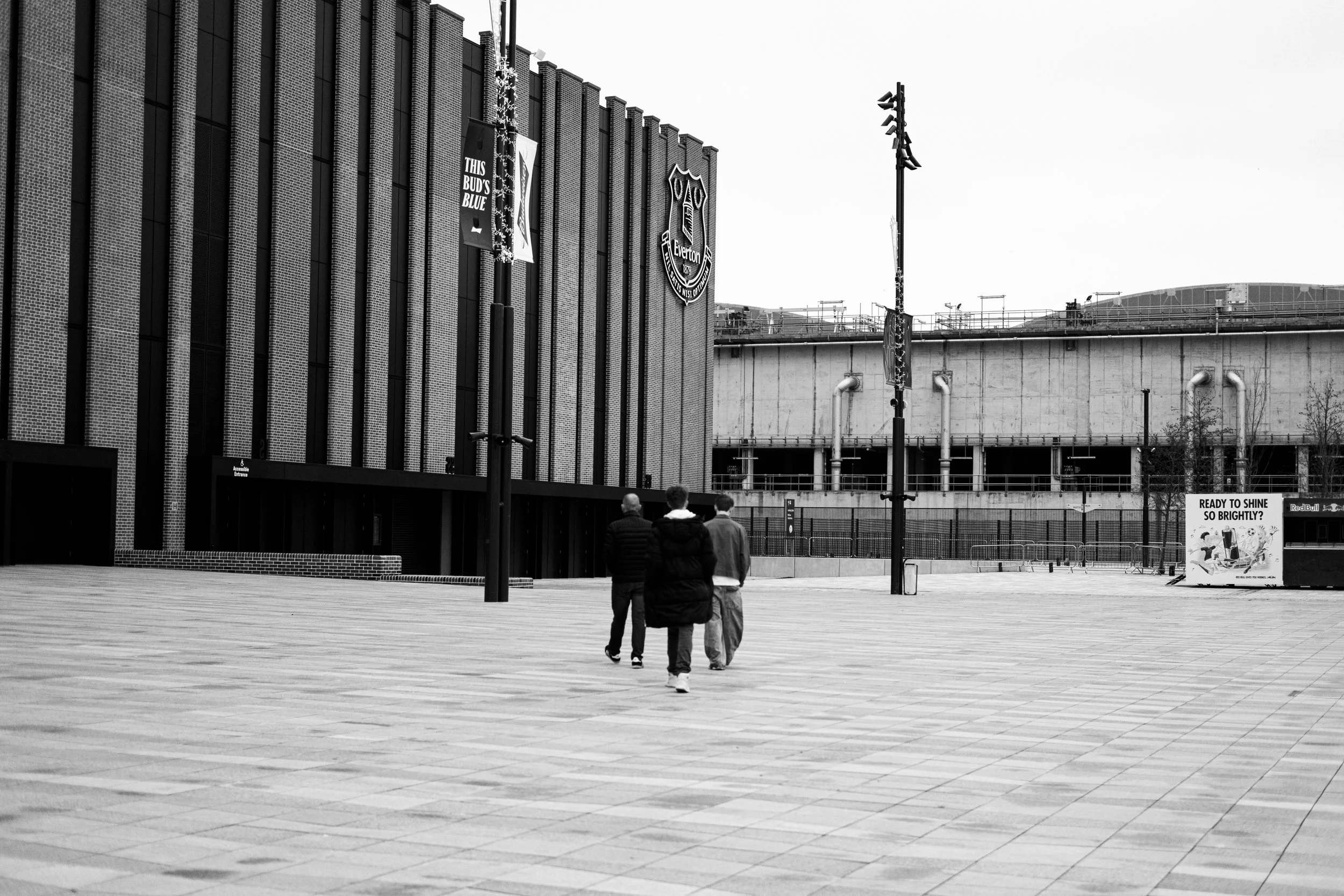 Three people walking in an open area outside a stadium with the Everton Football Club logo, surrounded by lamp posts, banners, and a large building.