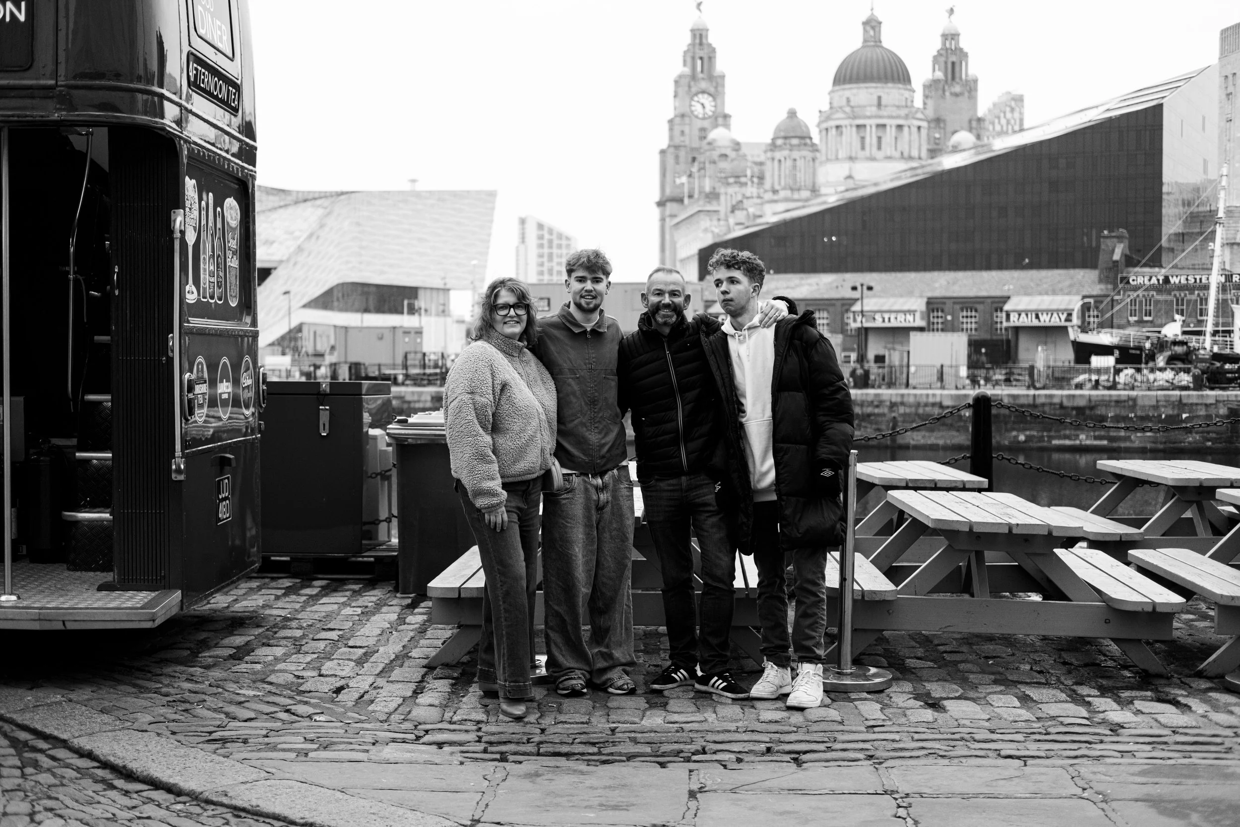Four people standing together outdoors near a waterfront with city buildings and dome in the background, posing for a photo. The scene is in black and white.