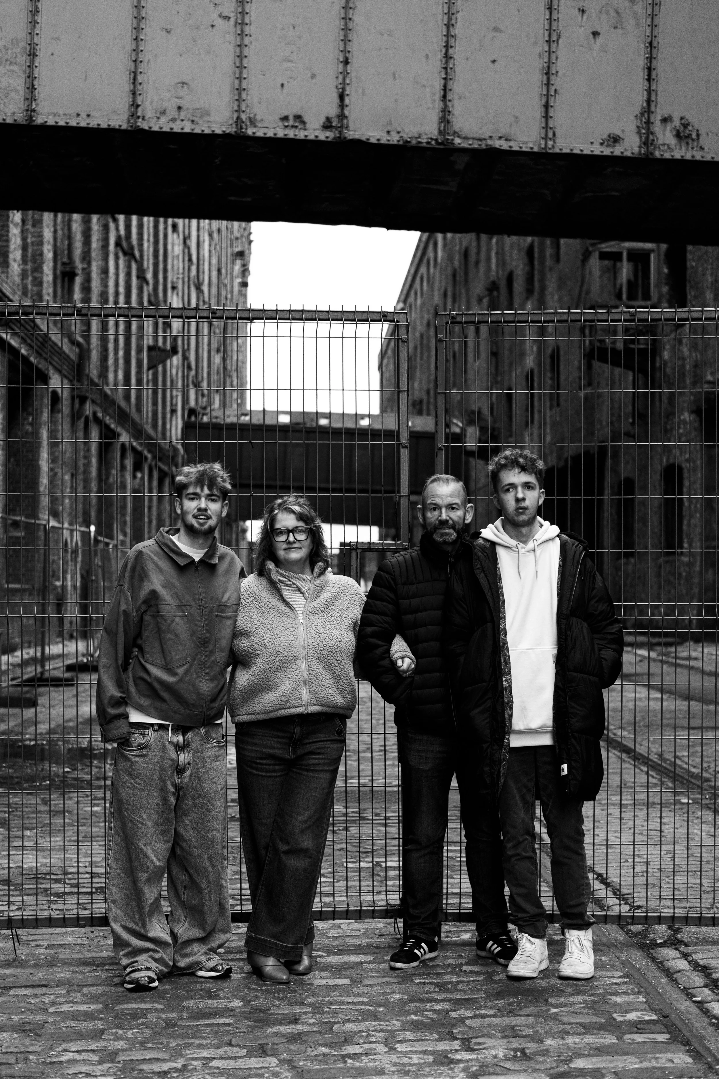 Four people standing behind a metal fence in an urban alleyway, black and white photo.
