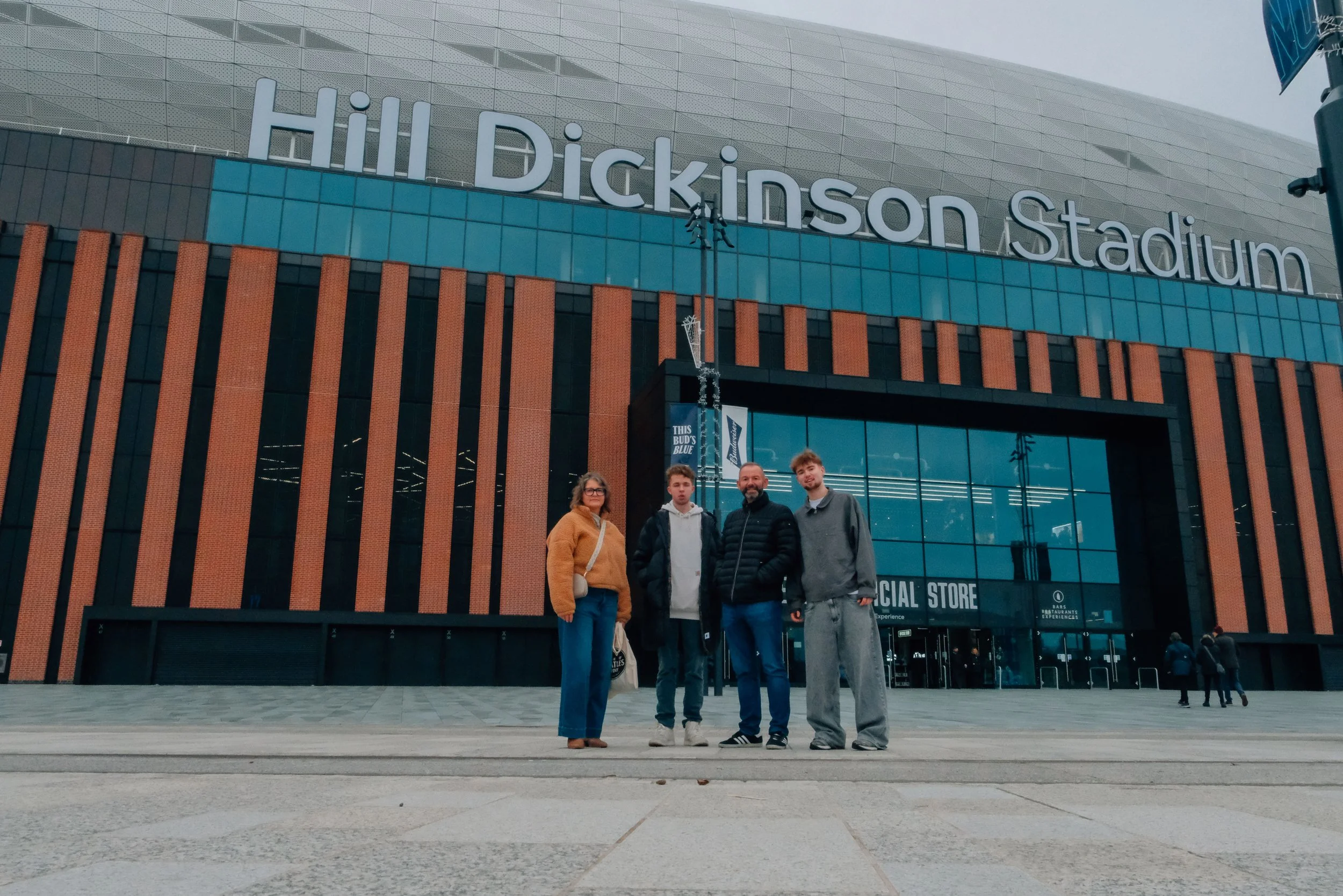 Four people standing in front of the Hill Dickinson Stadium, a large modern building with brick and glass facade, on a cloudy day.