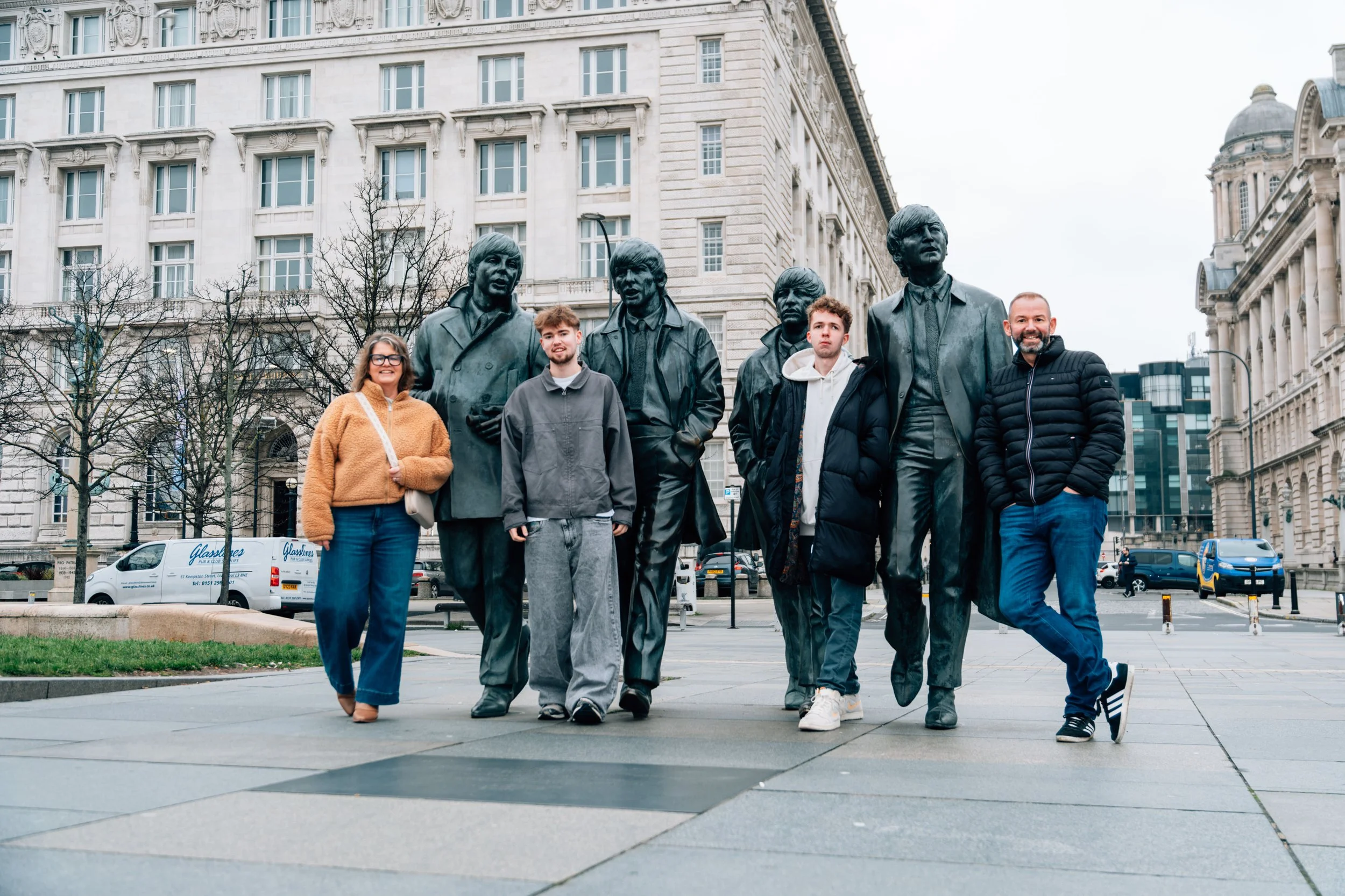 Four people standing in front of a statue of The Beatles in an urban setting with historic buildings in the background.